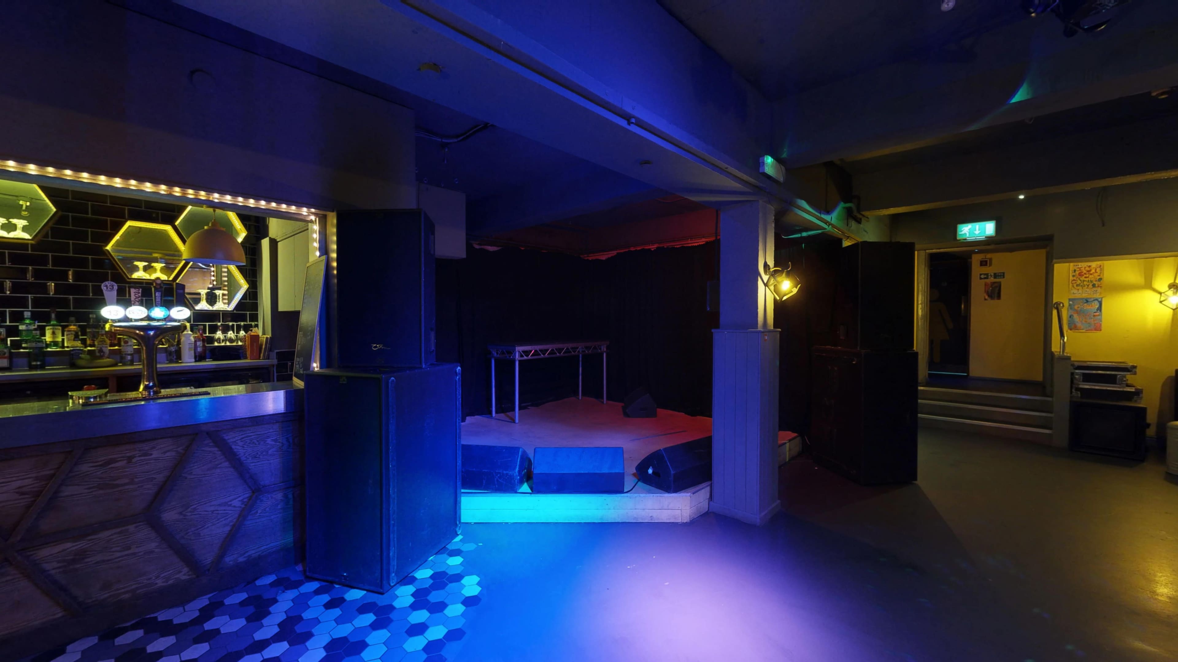 A dimly lit bar area with a performance stage in the back, featuring sound equipment and a seating area.