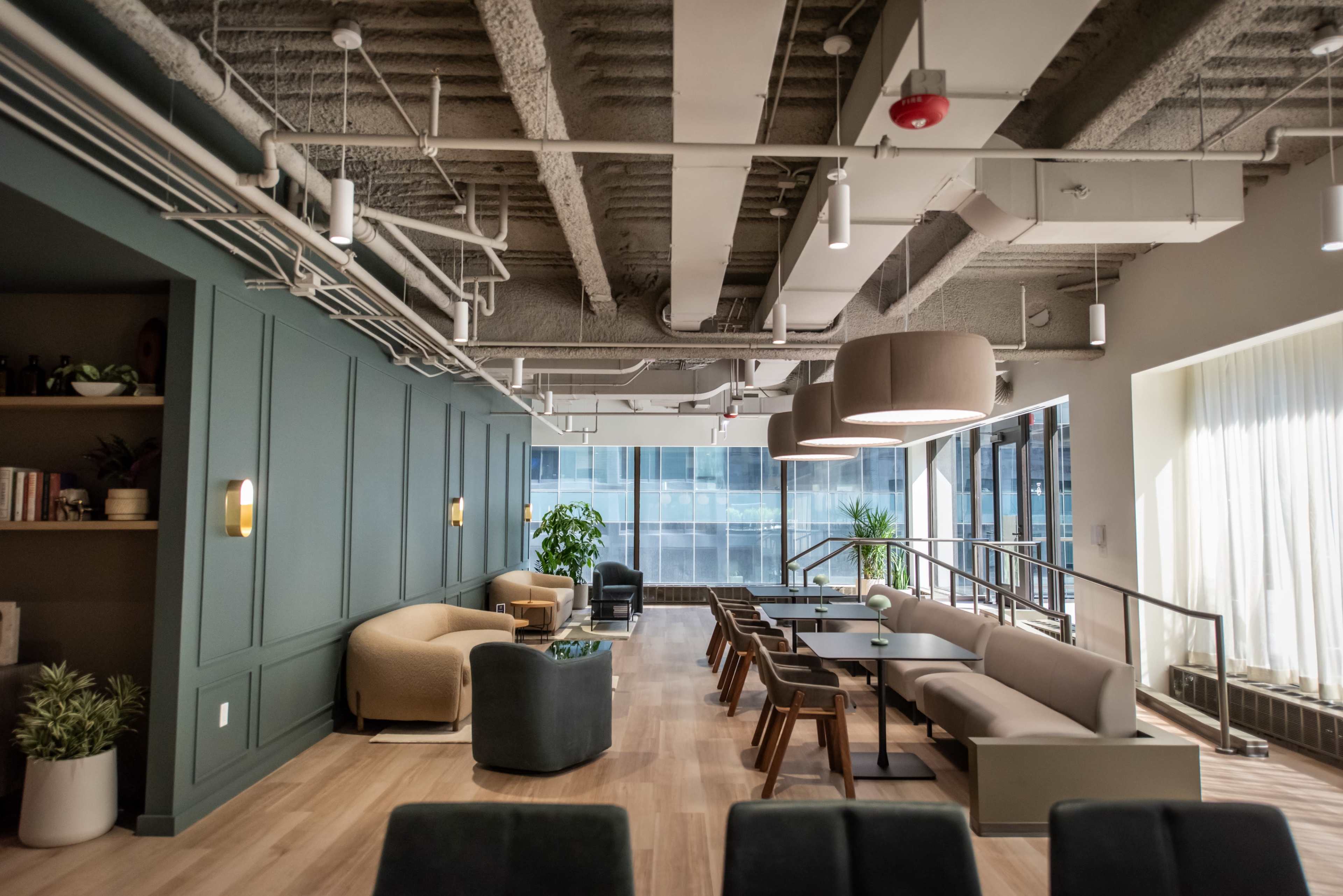 The image shows a modern office lounge area featuring various seating arrangements, a mix of light fixtures, and large windows providing natural light.