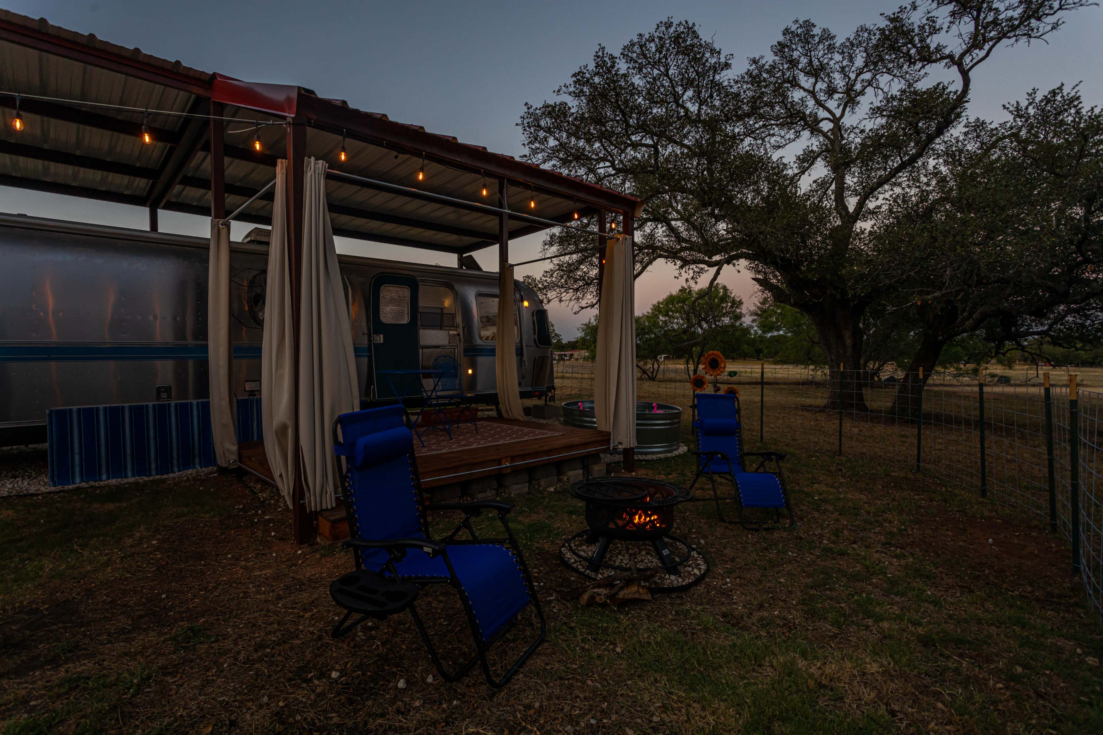 A cozy outdoor setup features a rustic trailer with a covered deck and a fire pit surrounded by portable chairs, set against a backdrop of trees at dusk.