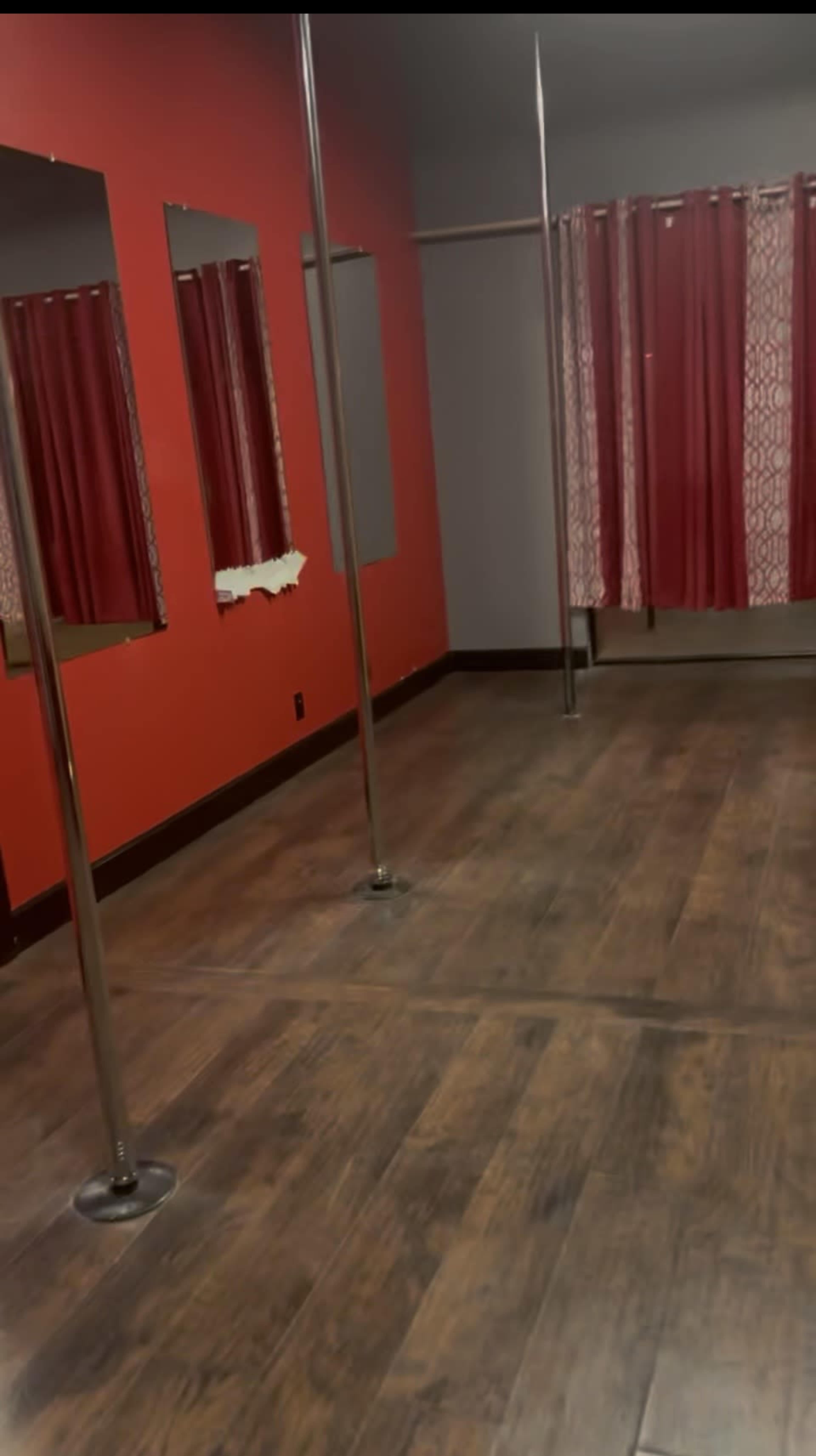 The image shows a dance studio with a wooden floor, red walls, and three metallic poles, along with curtains in the background.