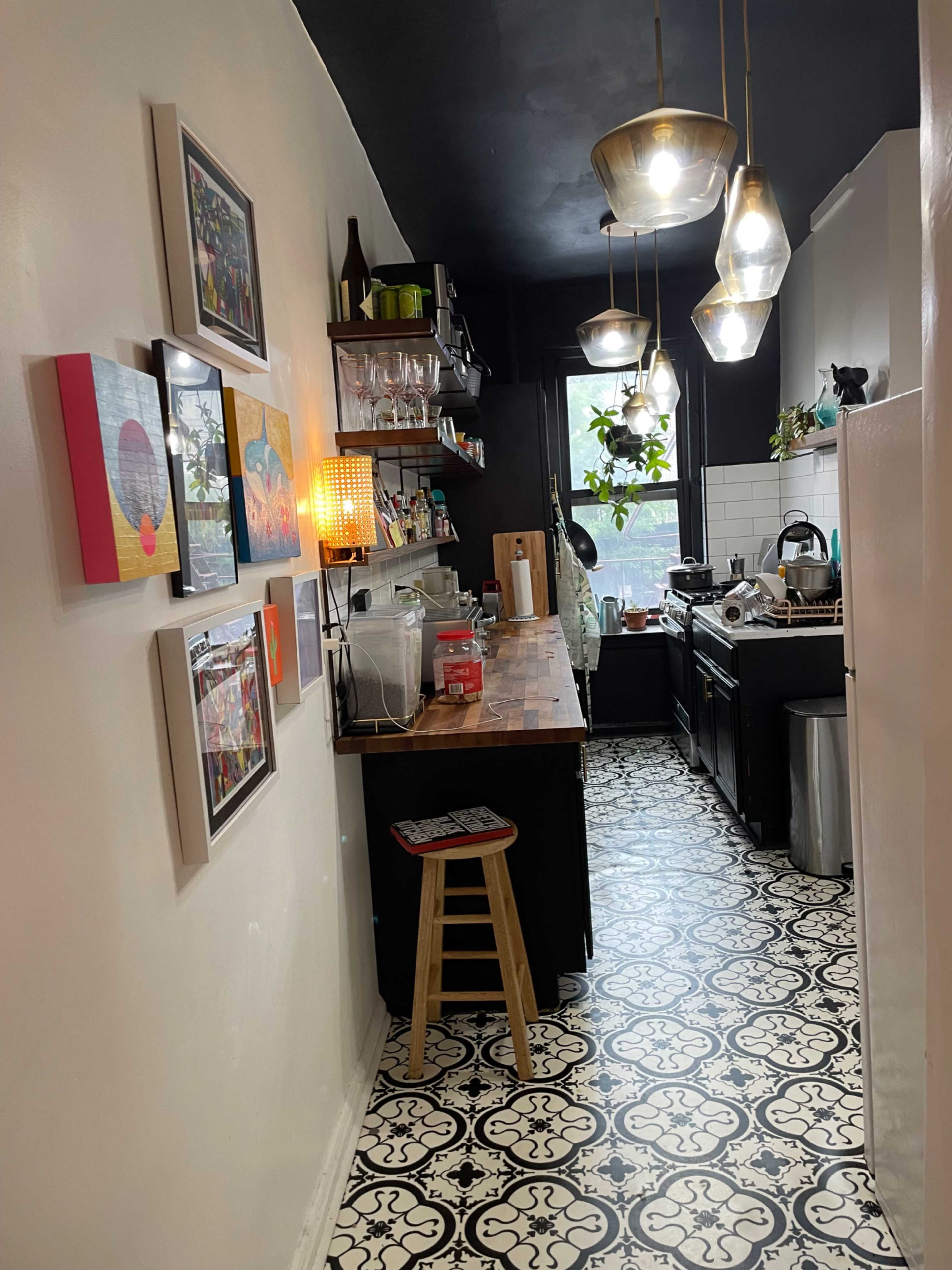 A narrow kitchen with black and white patterned flooring, shelves filled with various items, and a bar-style seating area with a single stool.