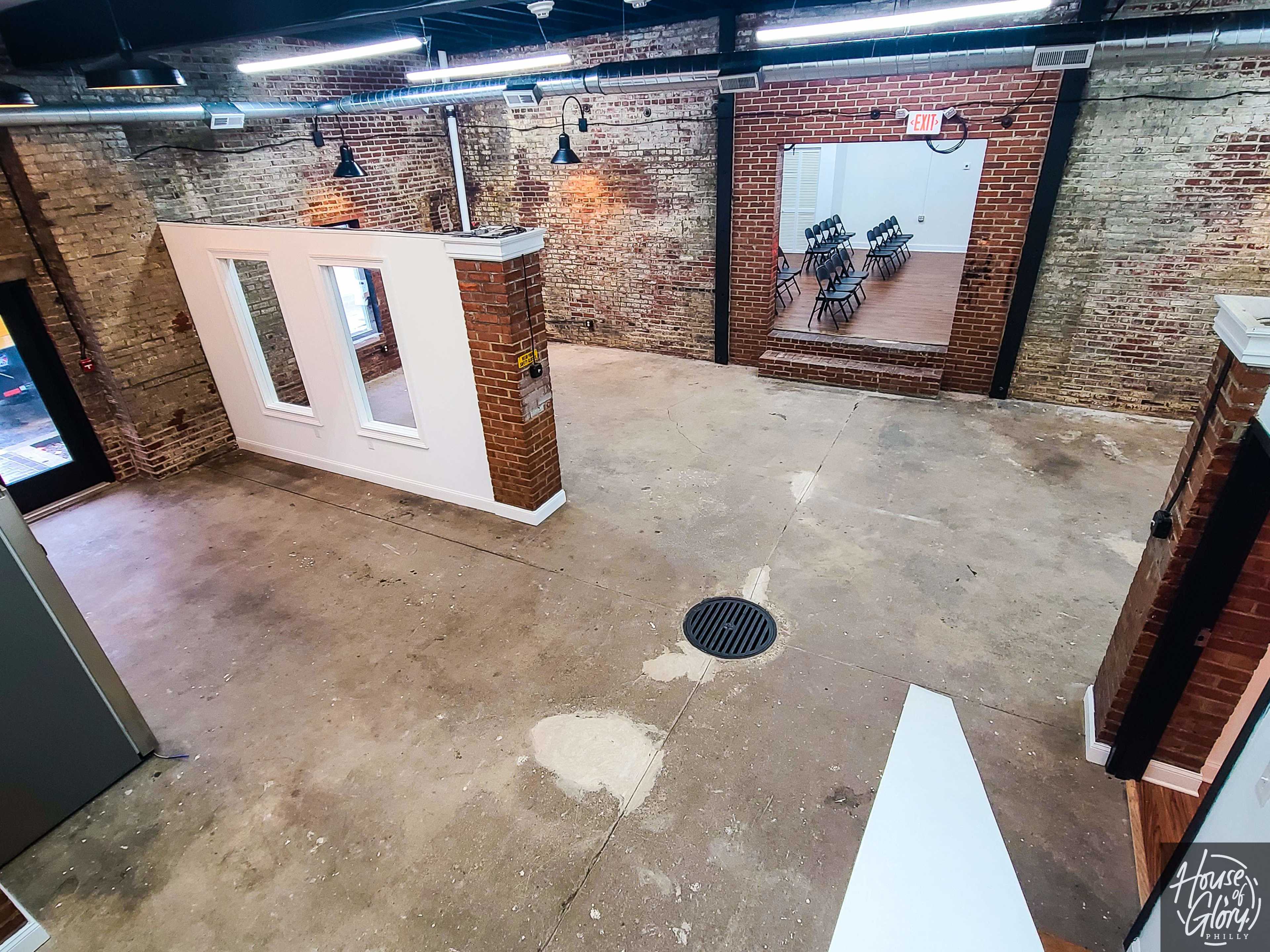 A spacious, unfinished interior with exposed brick walls and a large open area, featuring two windows and a doorway leading to a room with rows of chairs.