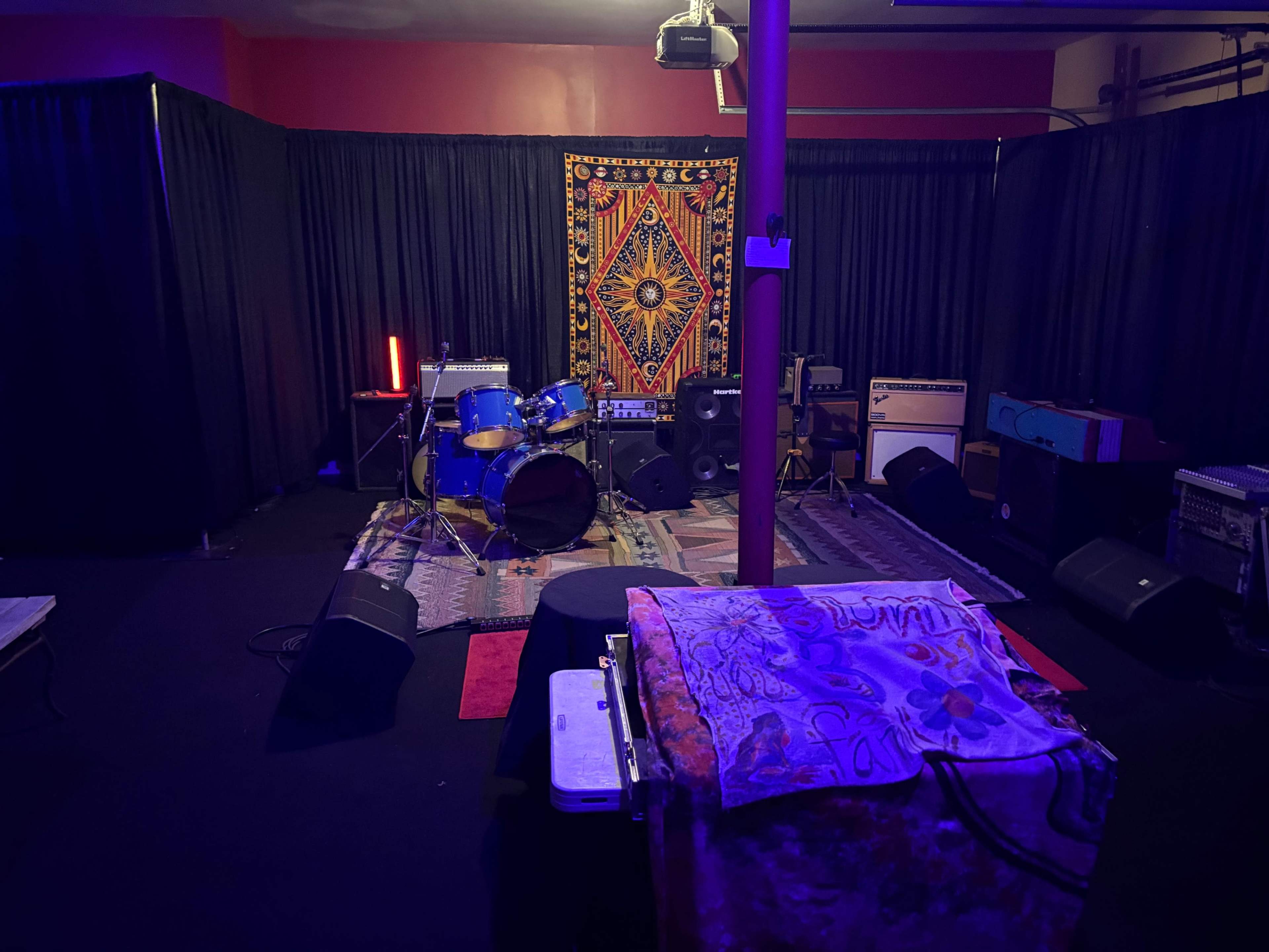 The image shows a dimly lit rehearsal space with a drum set, amplifiers, and a colorful tapestry hanging on the back wall.