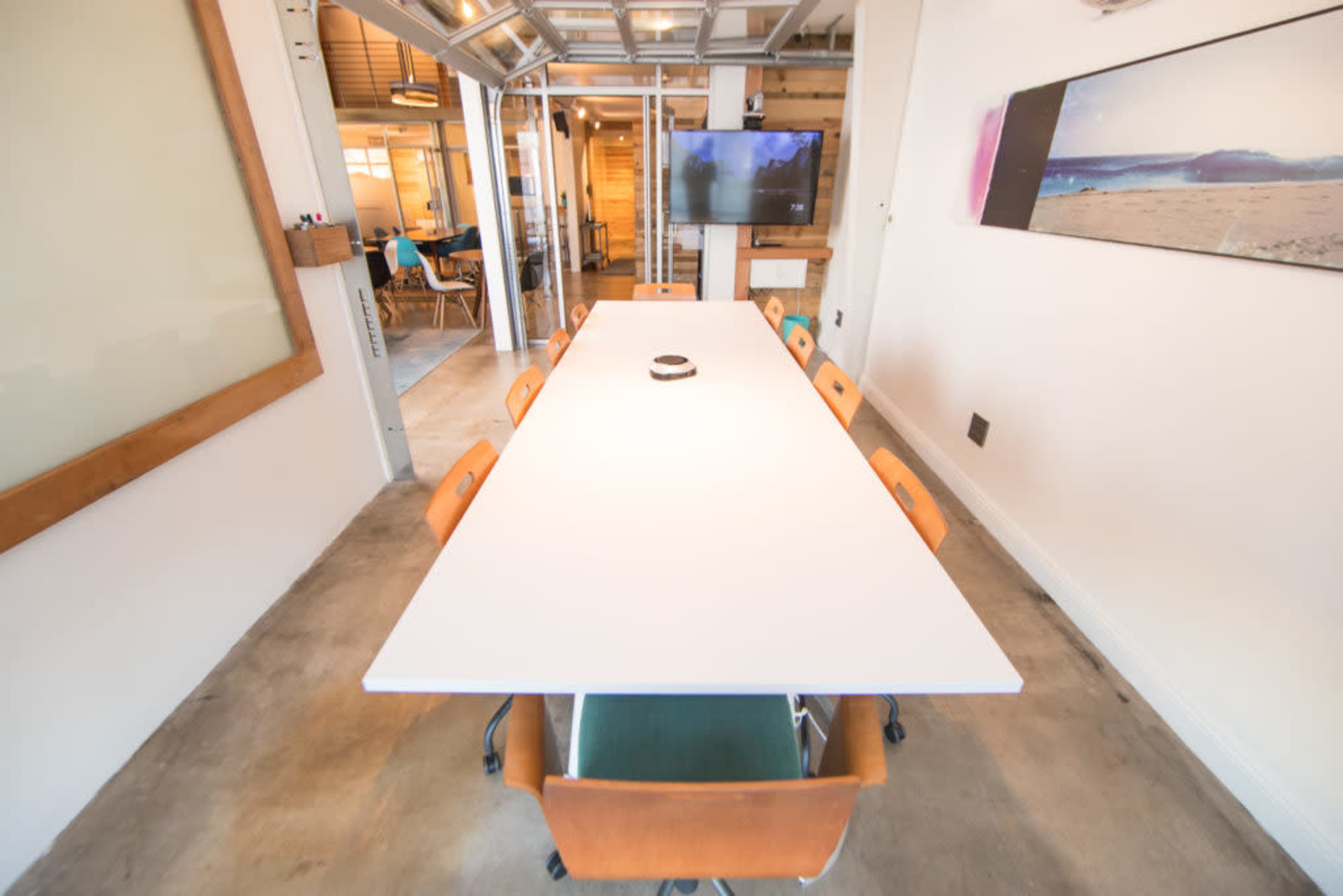 A long, white conference table is set with chairs around it in a modern office space, with a television and additional seating visible in the background.