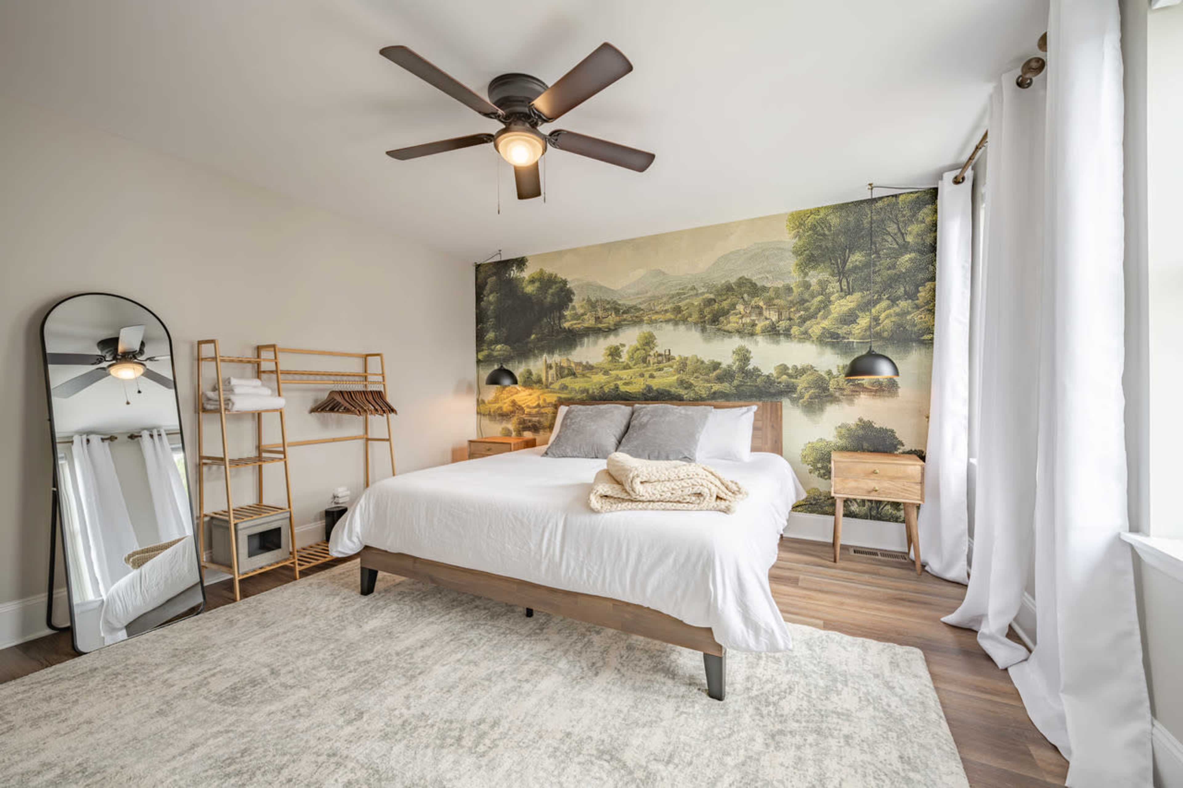 A bedroom features a queen-sized bed with a scenic mural of a landscape, complemented by a ceiling fan and minimalist decor.