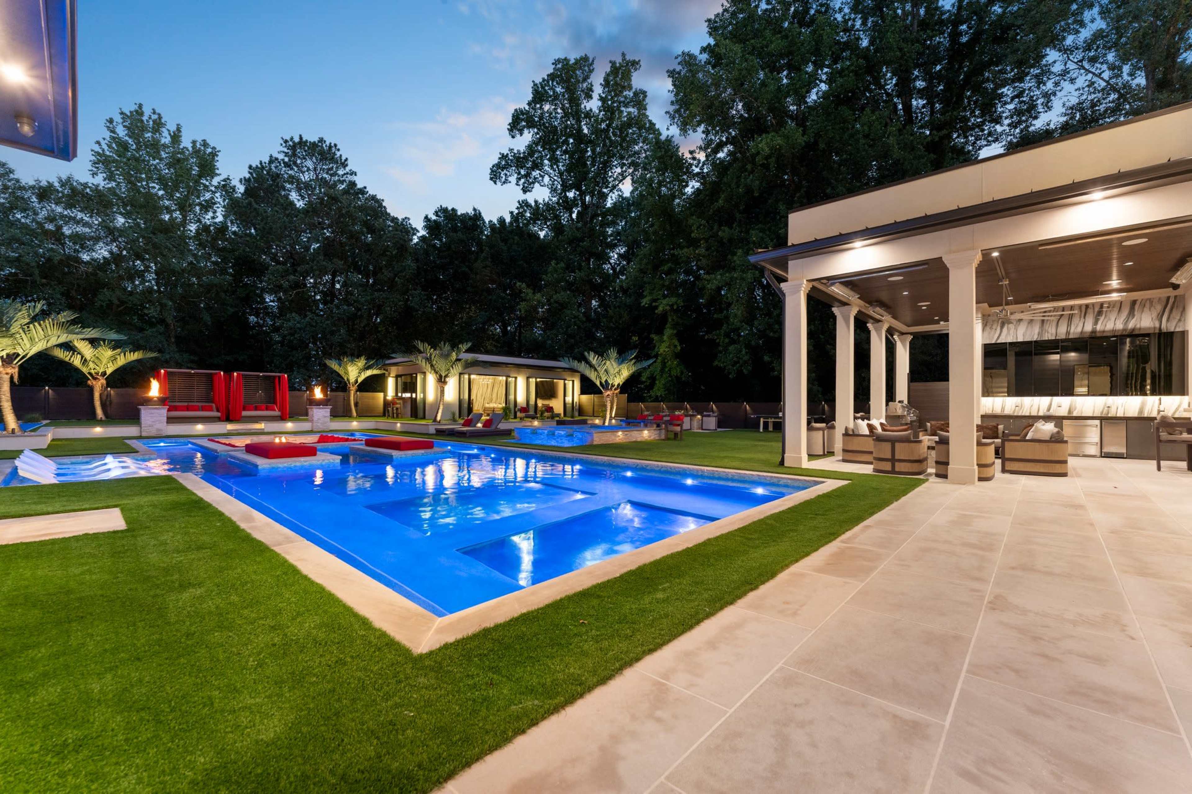 The image shows a modern backyard featuring a well-lit swimming pool surrounded by manicured grass, lounge seating, and palm trees.