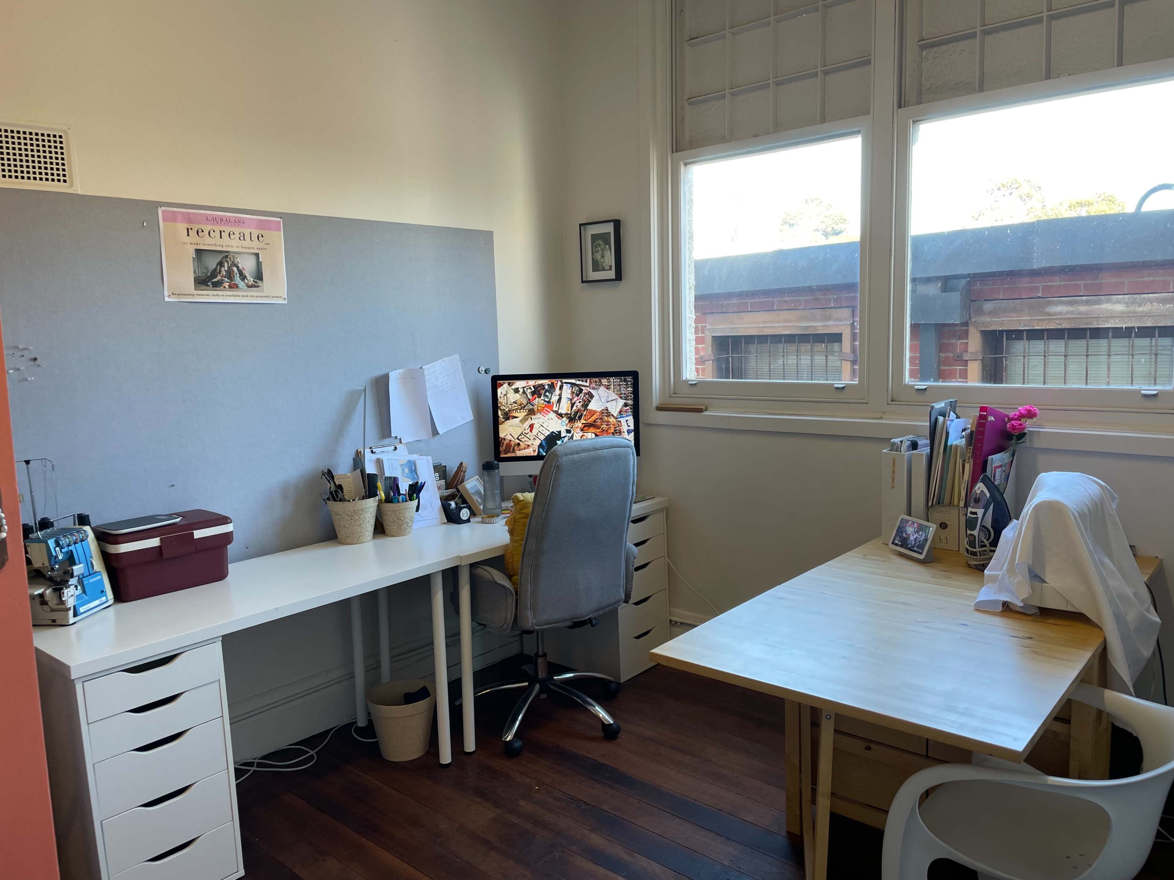 The image shows a brightly lit home office with a desk, an ergonomic chair, a computer monitor displaying a colorful screensaver, and another table with a few personal items.