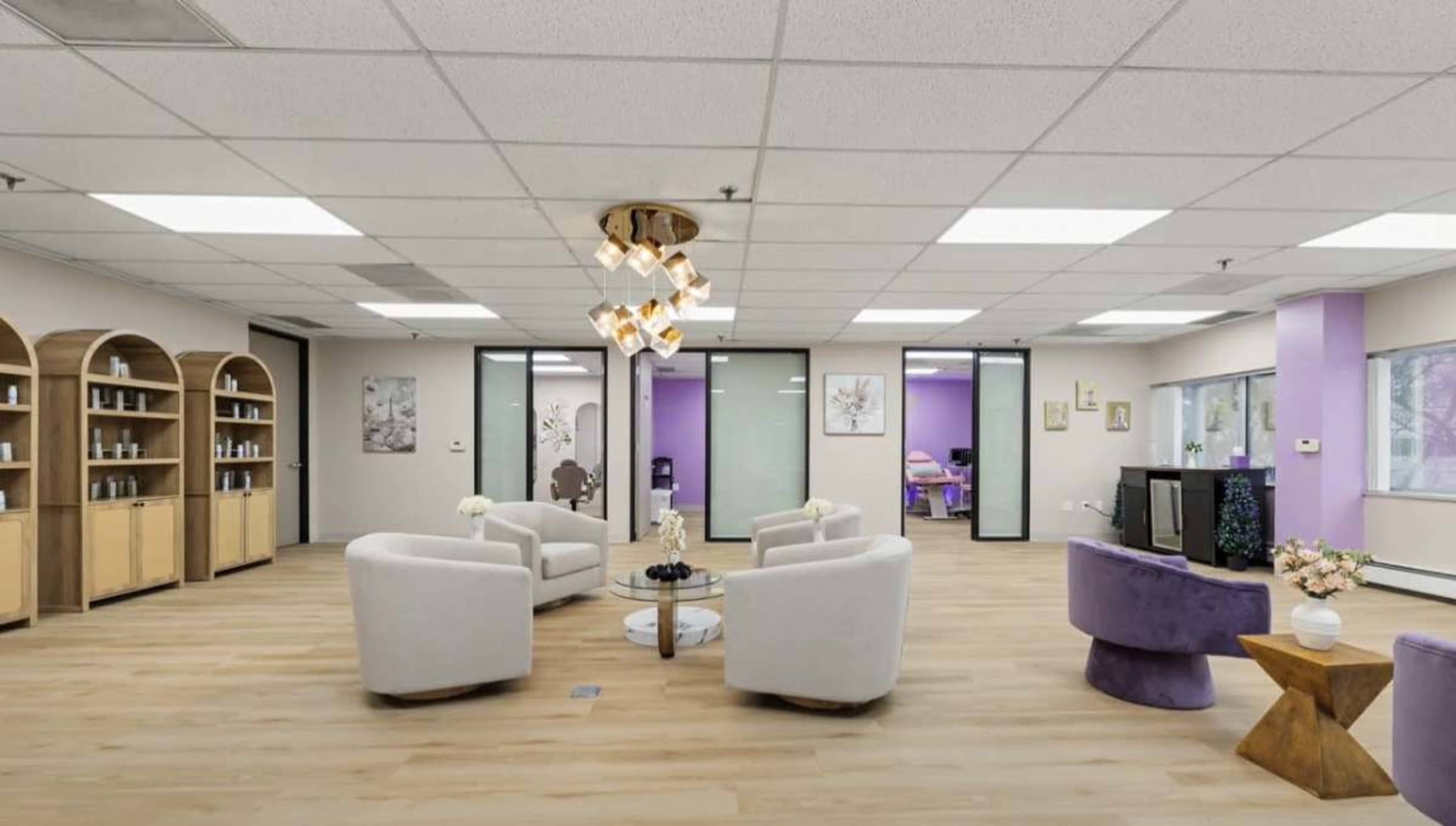 The image shows a modern, open office space featuring light-colored wooden flooring, white chairs arranged around a small table, and purplish walls in the background.