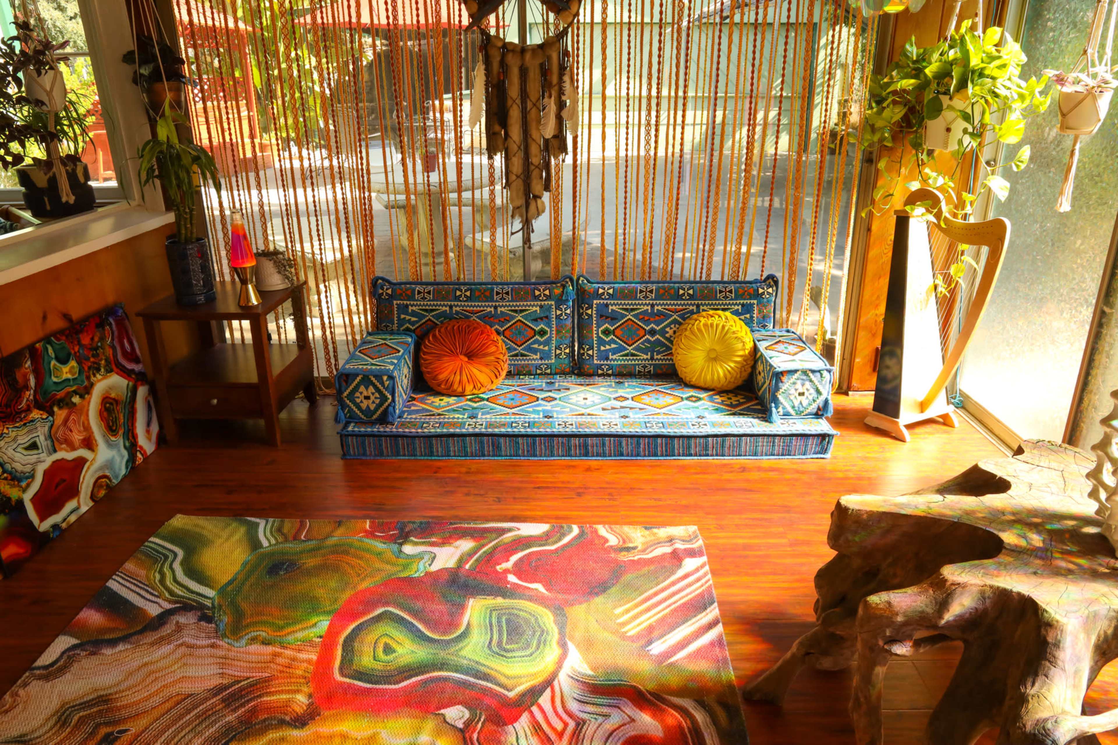 A brightly decorated interior with a patterned couch, colorful pillows, and a vibrant area rug, surrounded by plants and natural light.