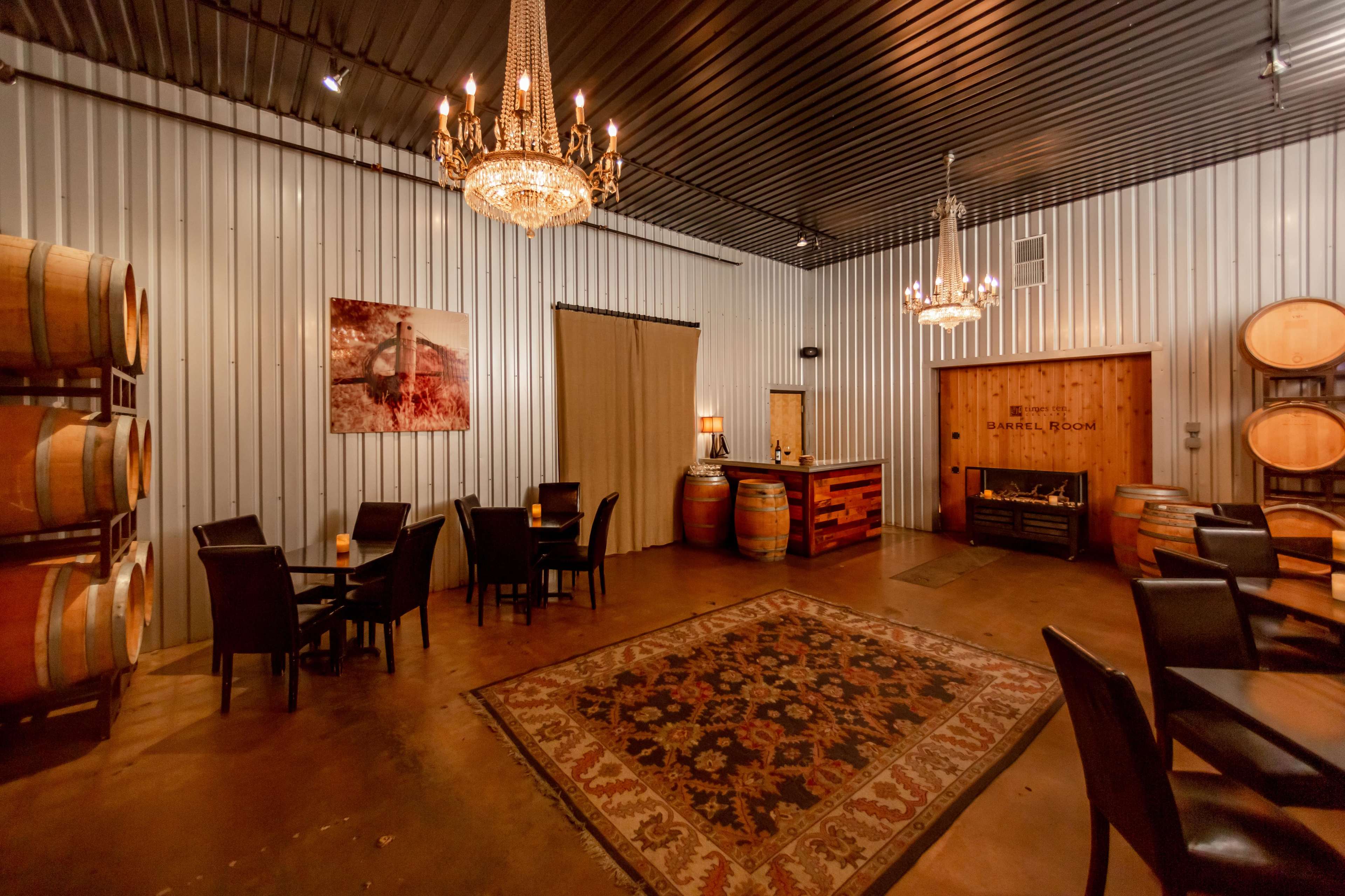 A wine tasting room featuring wooden barrels, a small bar, and a decorative rug under a set of chandeliers.