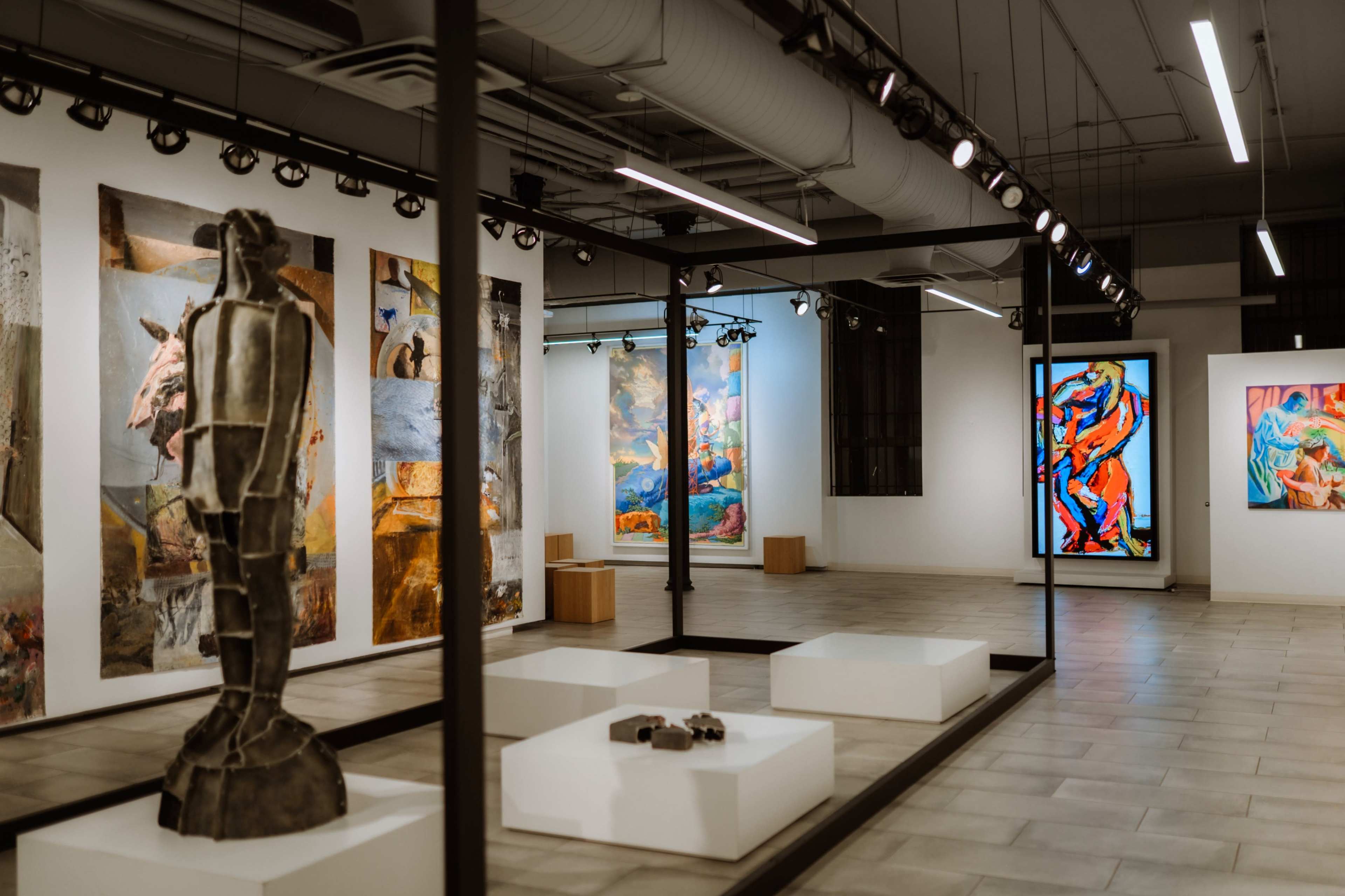 The image shows an art gallery featuring various colorful paintings on white walls, with a sculpture on a pedestal in the foreground and a modern lighting setup above.