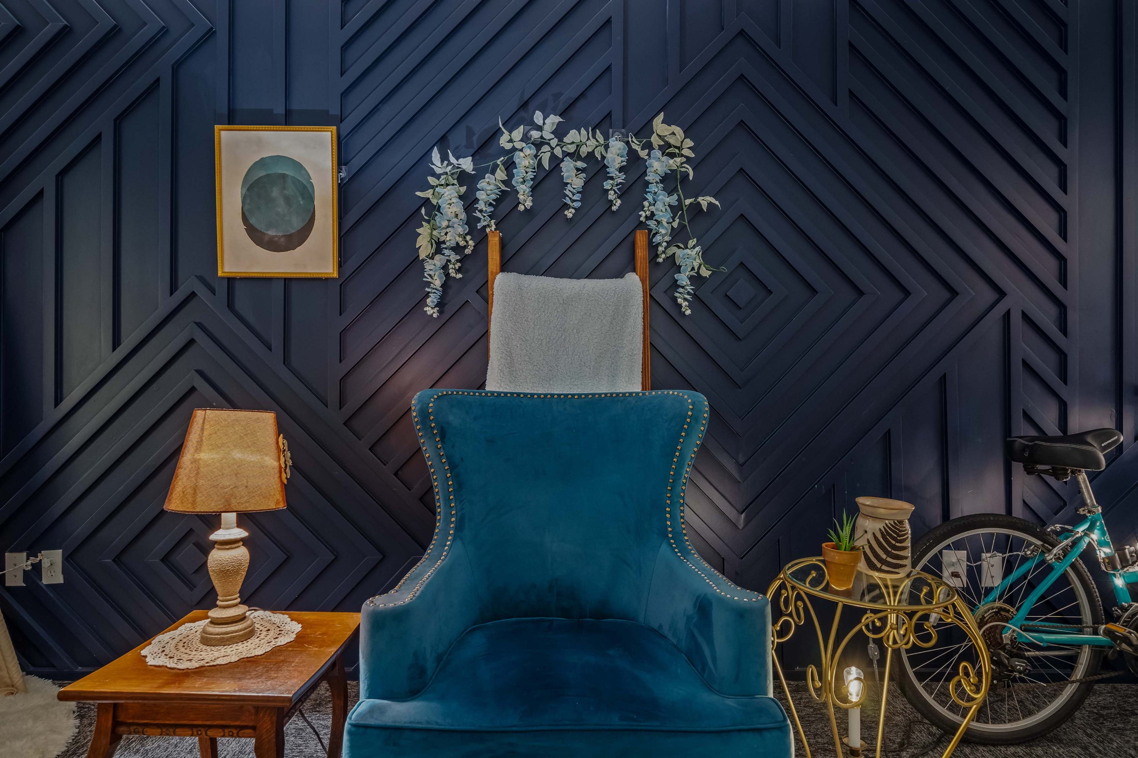 A blue velvet armchair is positioned near a patterned wall, accompanied by a lamp, a small table, and a bicycle in the background.