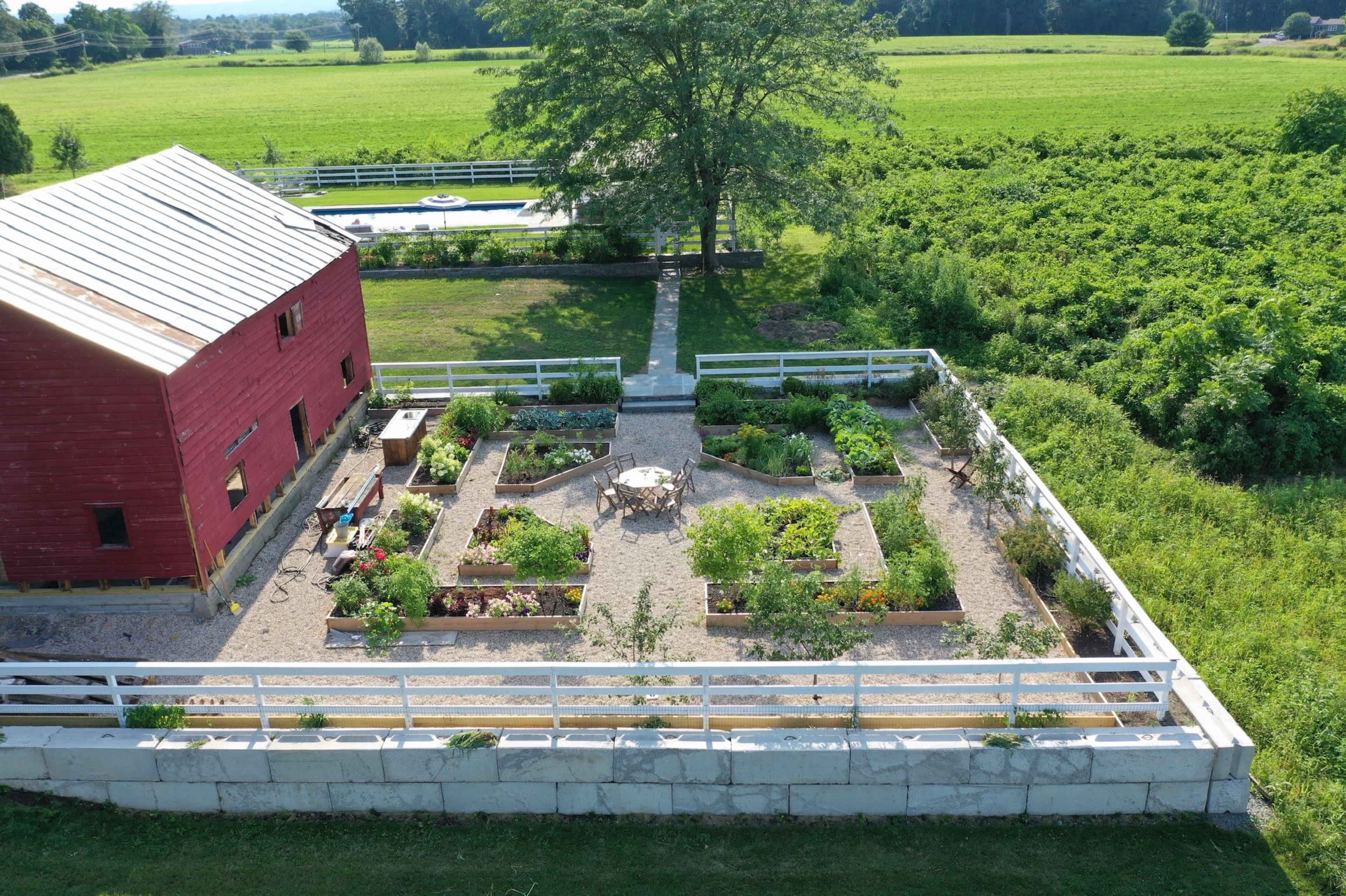 A large garden arranged in a geometric layout with various plants, bordered by a red barn and a grassy area, alongside a pool in the background.