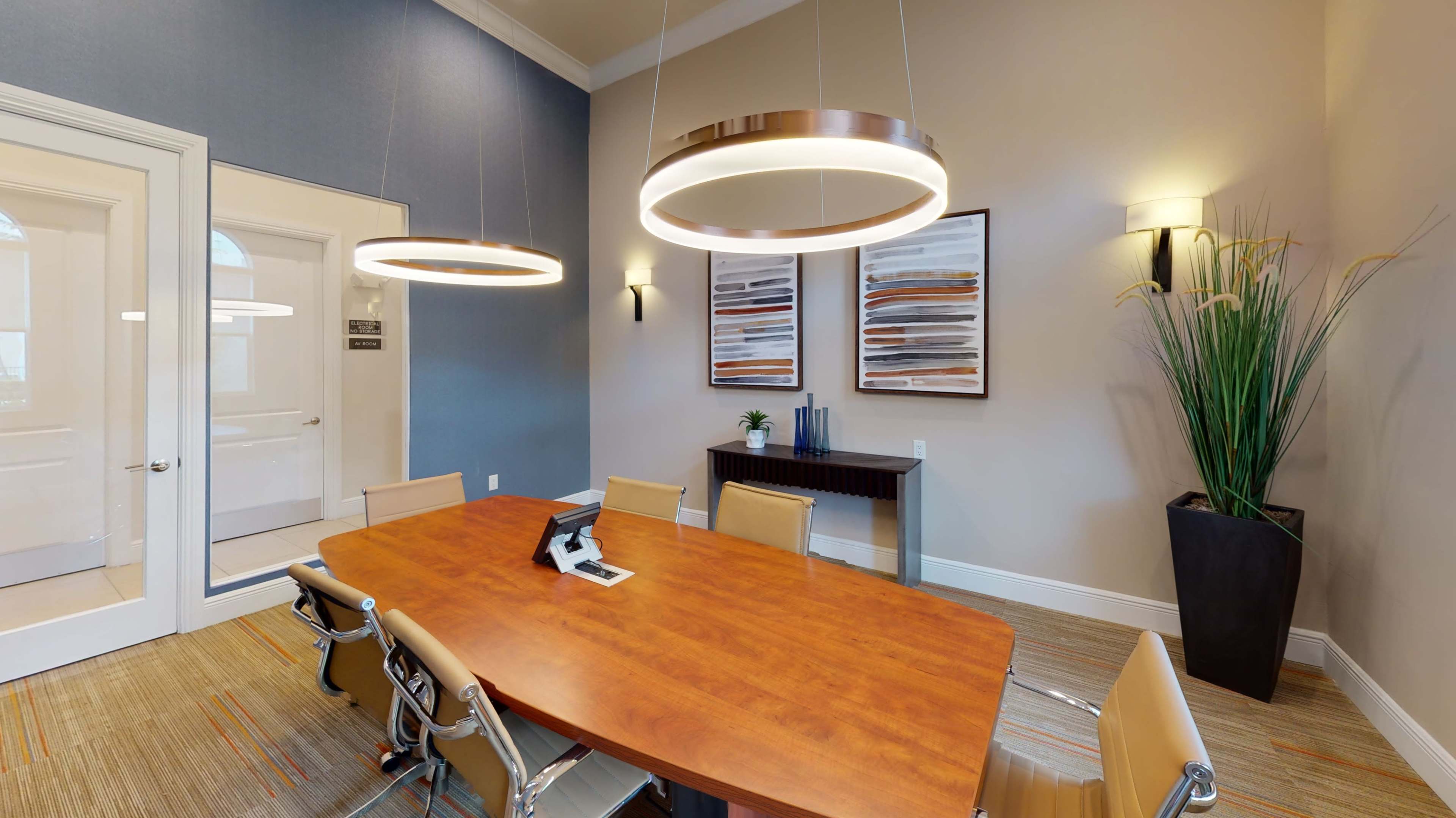 A modern conference room features a large wooden table surrounded by leather chairs, with two circular pendant lights hanging above and abstract art on the walls.