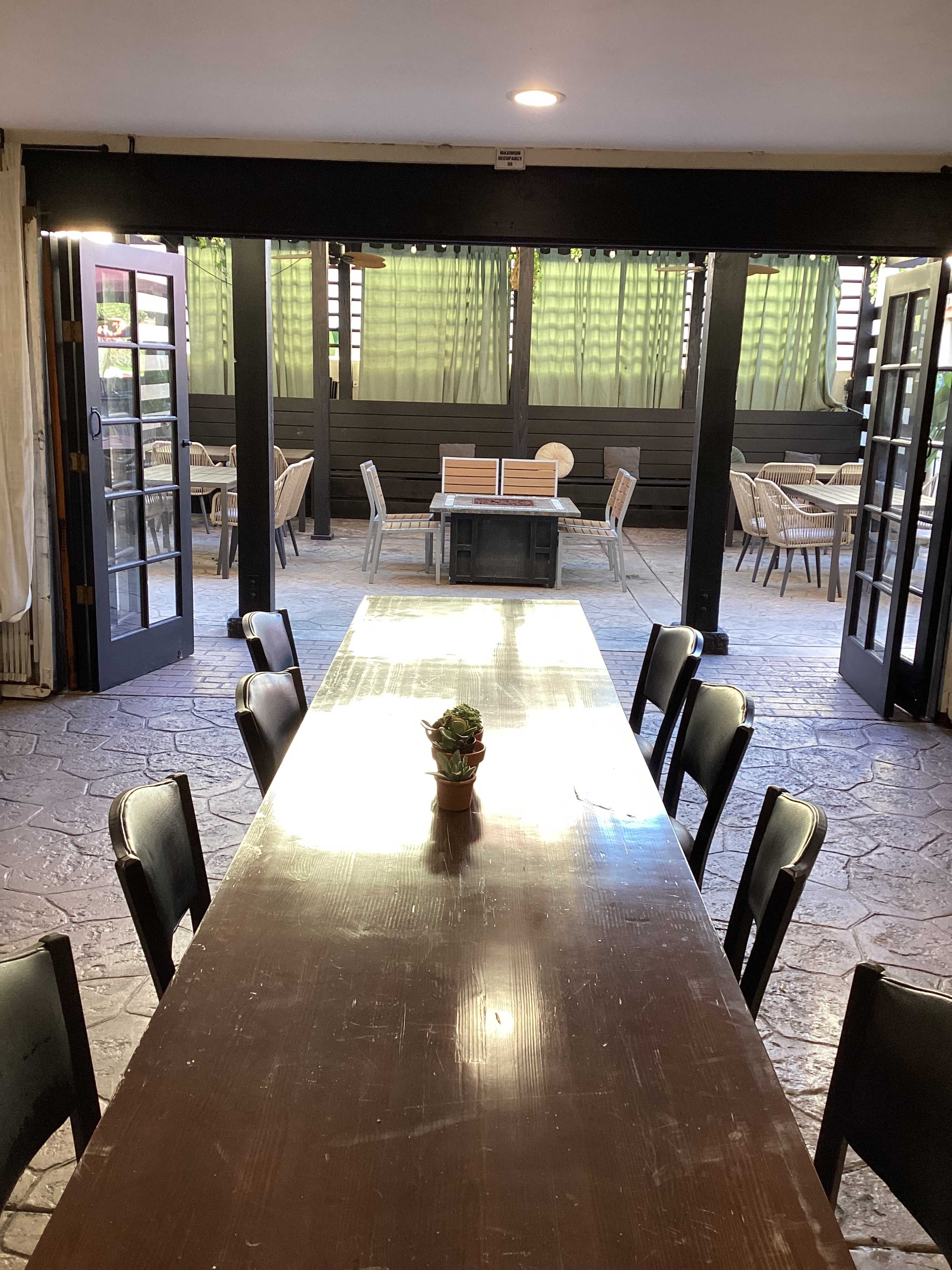 A long wooden table with a small plant centerpiece sits in a spacious dining area, flanked by several empty chairs and large open doors leading to an outdoor seating area.