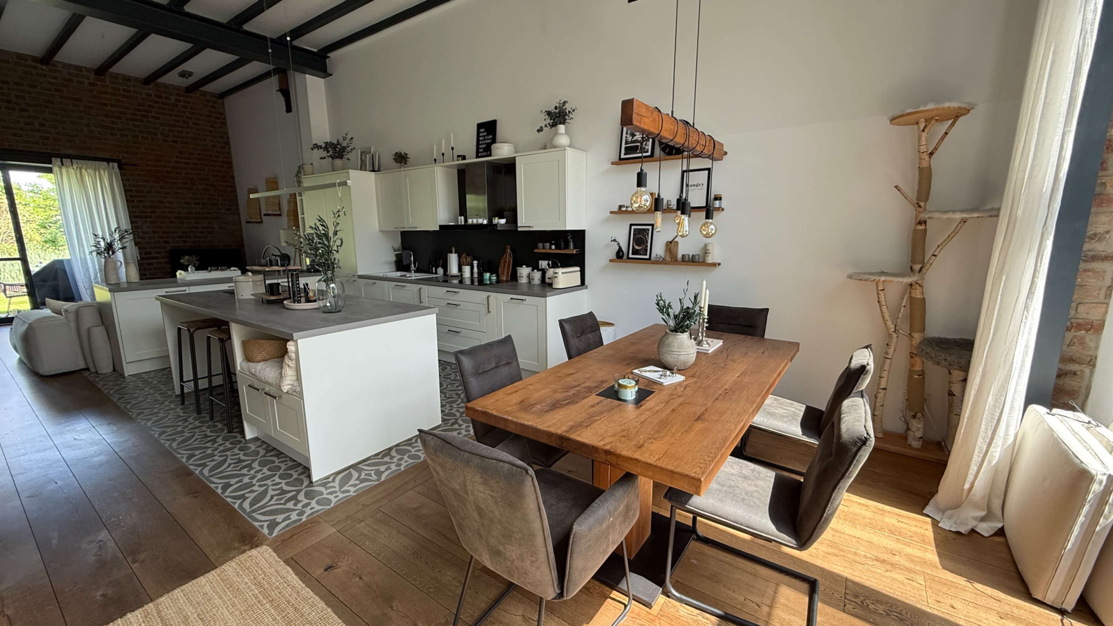 The image shows a modern dining area with a wooden table surrounded by chairs, an adjacent kitchen featuring white cabinets, and a cozy seating area with large windows.