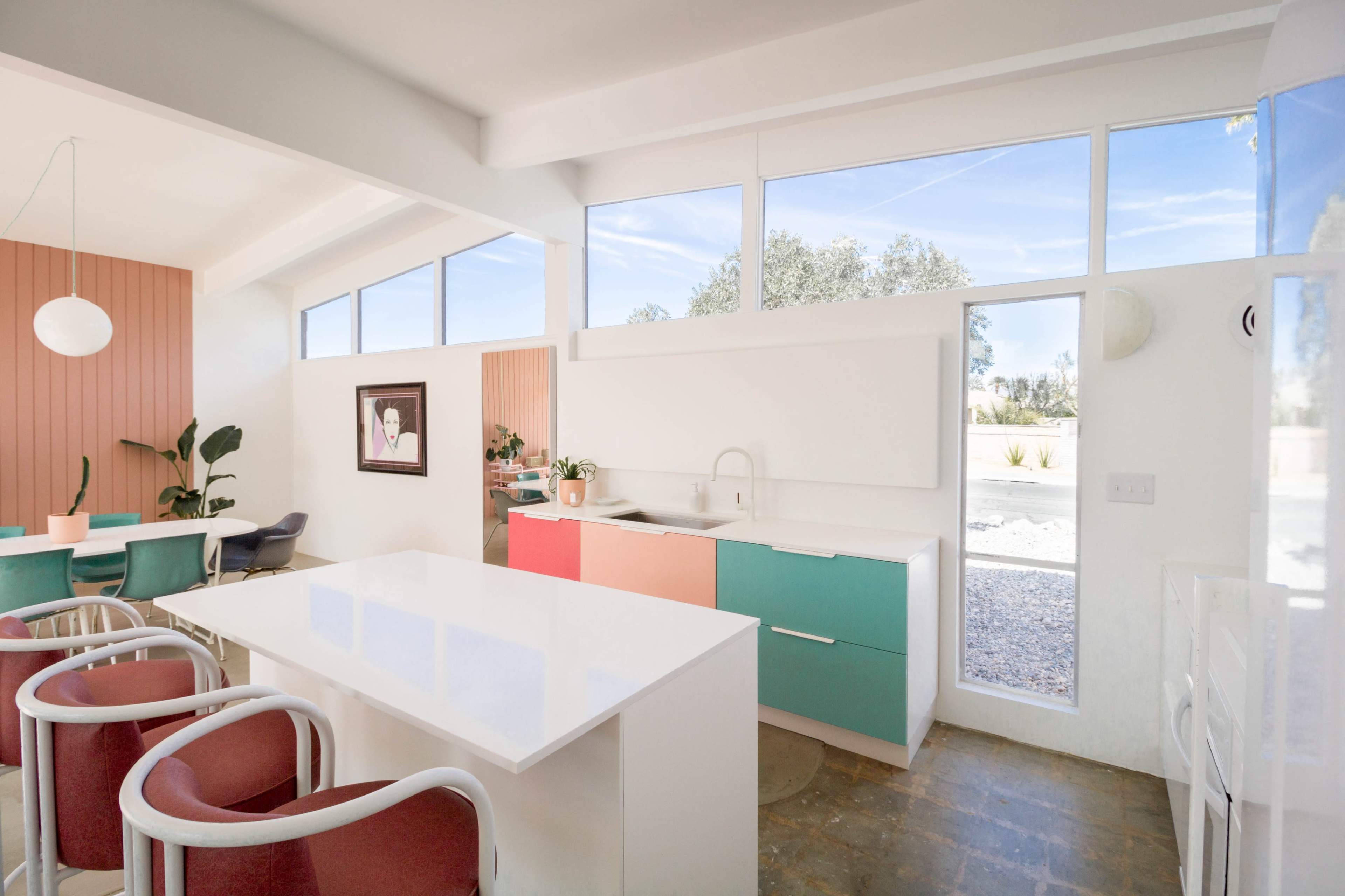 A modern kitchen features colorful cabinets, a sleek white countertop, and large windows that allow natural light to fill the space.