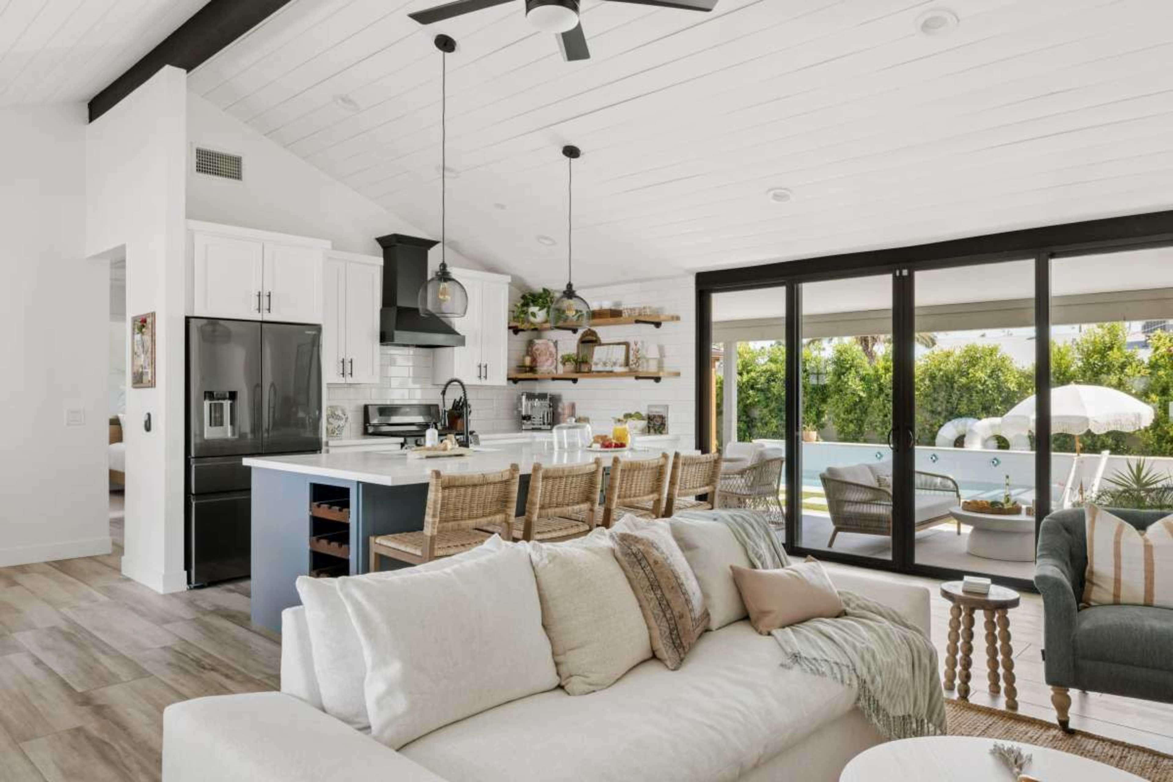 The image shows a modern open-concept living area with a kitchen featuring a large island, a dining area with bar stools, and sliding glass doors leading to an outdoor patio with greenery.