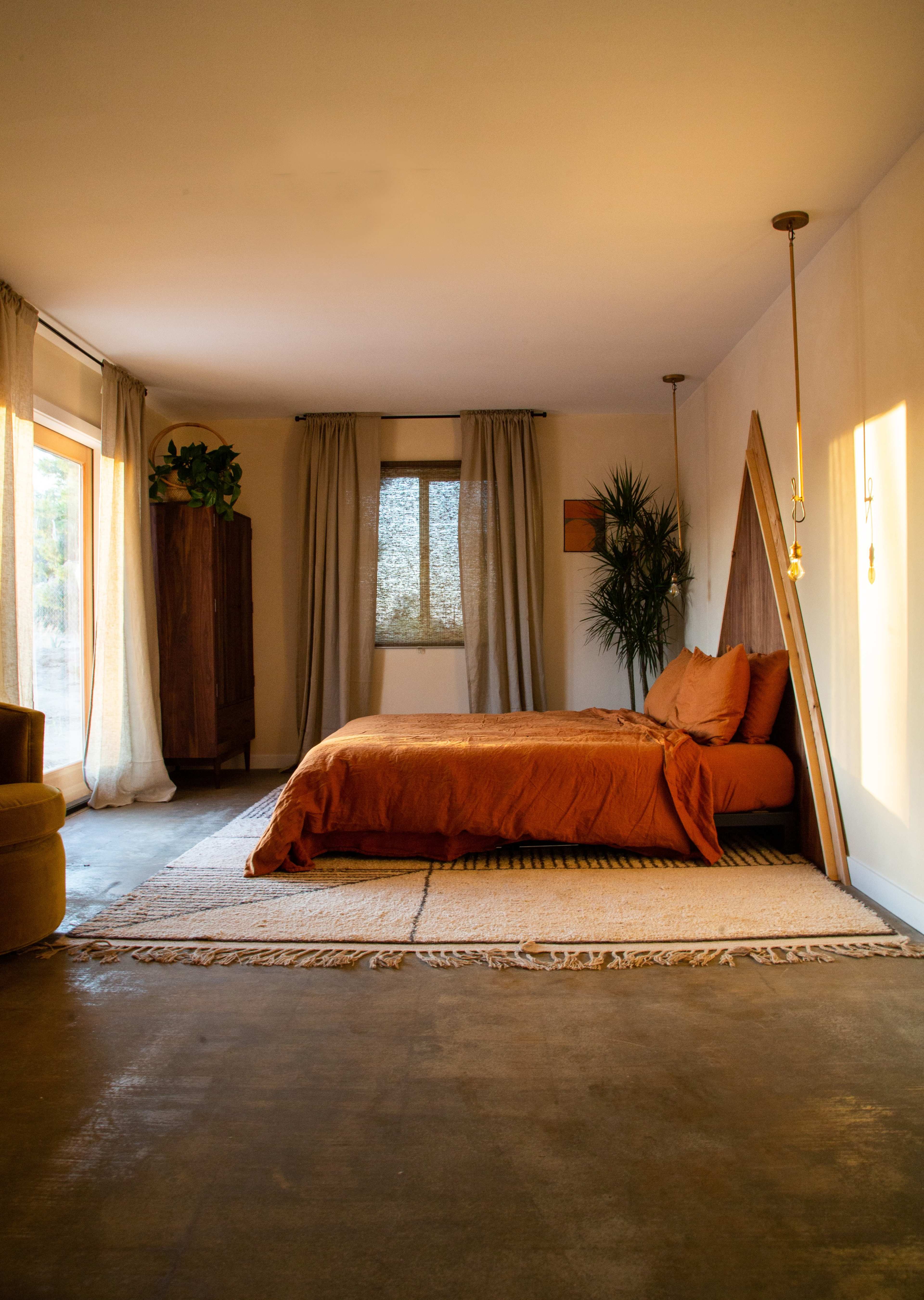 The image shows a spacious bedroom featuring an orange bedspread, large windows with sheer curtains, and a potted plant beside the bed.