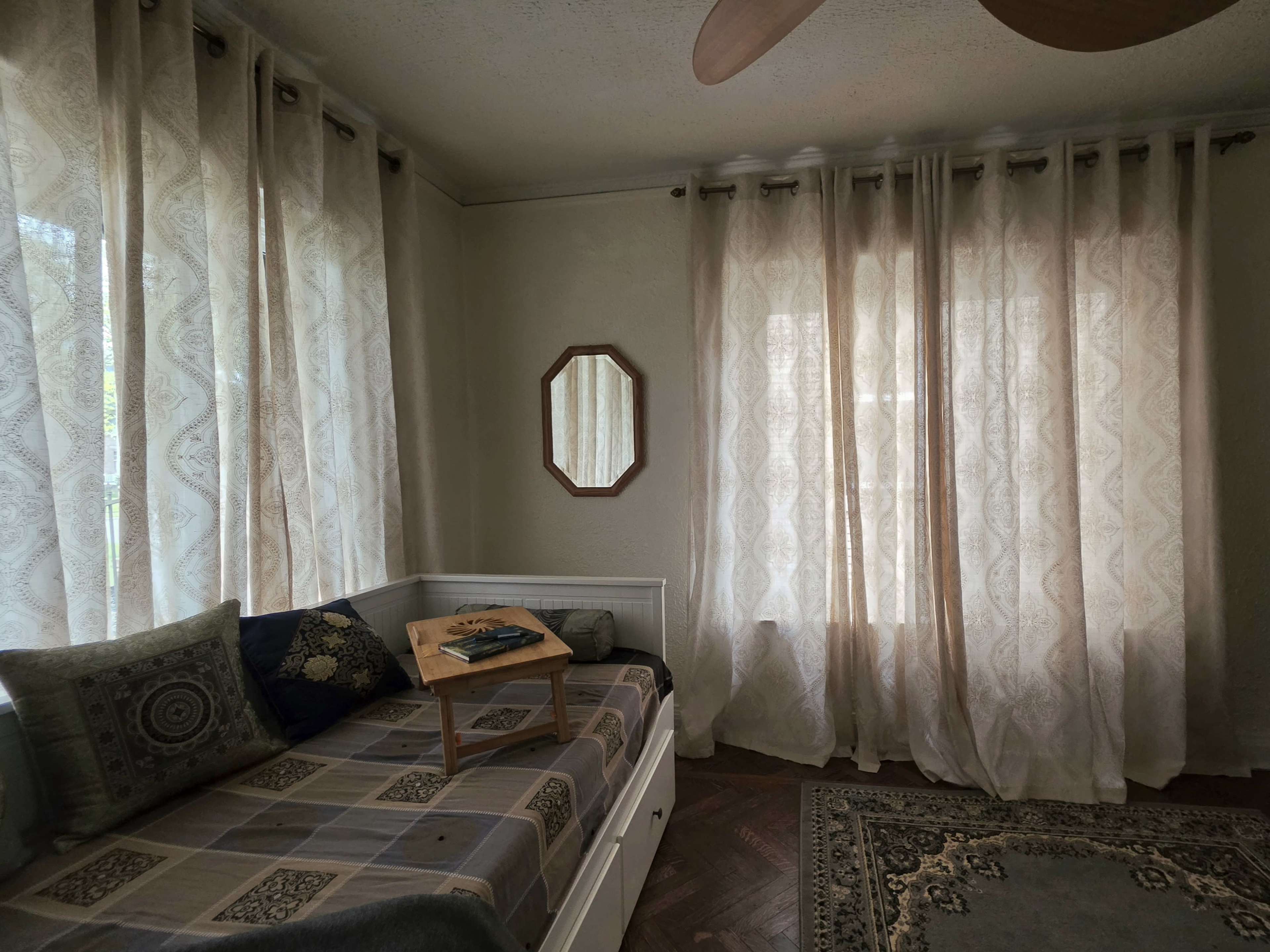 The image shows a light-filled room featuring a daybed with patterned bedding, a wooden tray on top, and sheer curtains framing the windows.