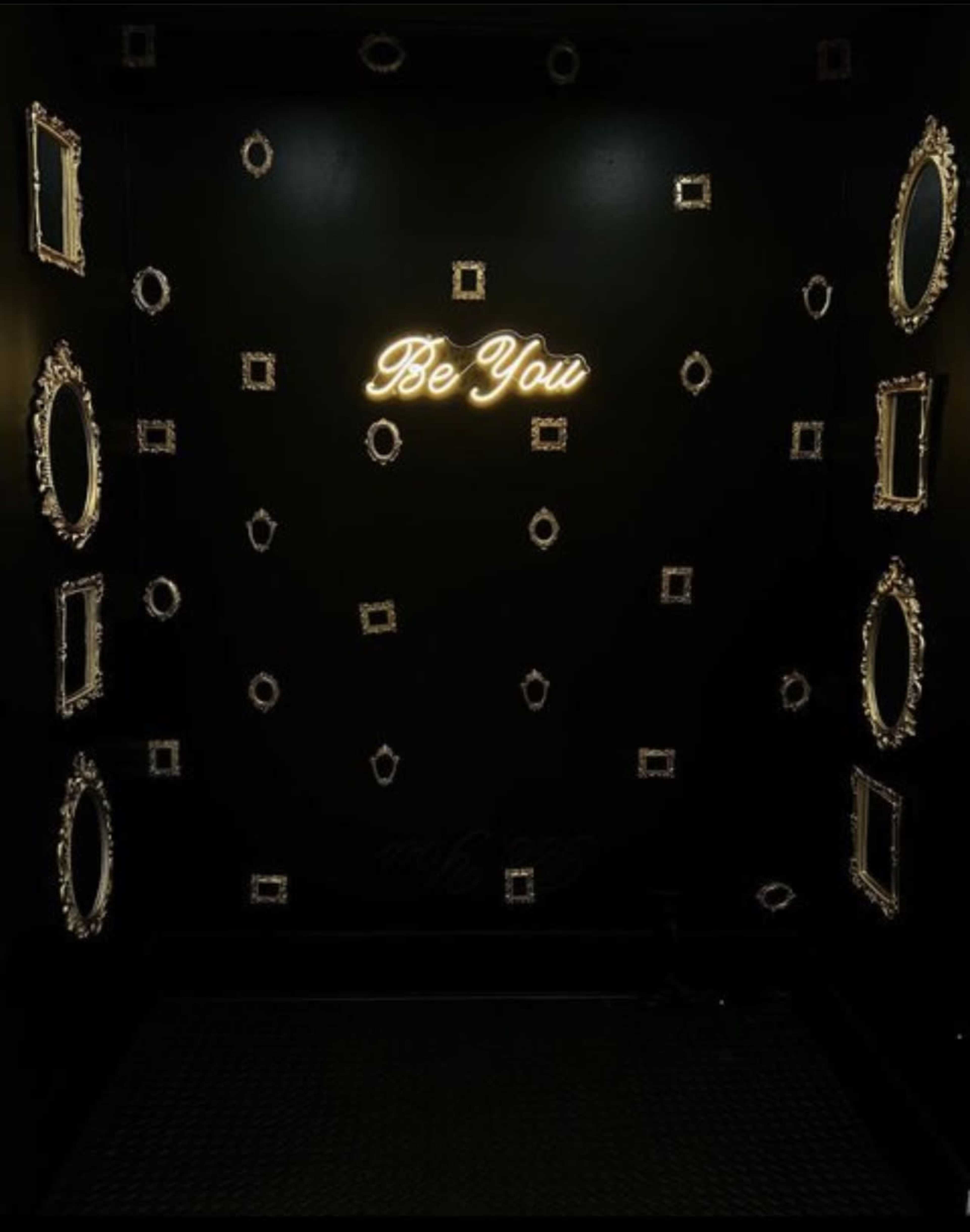 A dark room with multiple ornate mirrors on the walls and a neon sign that says "Be You" illuminated in the center.