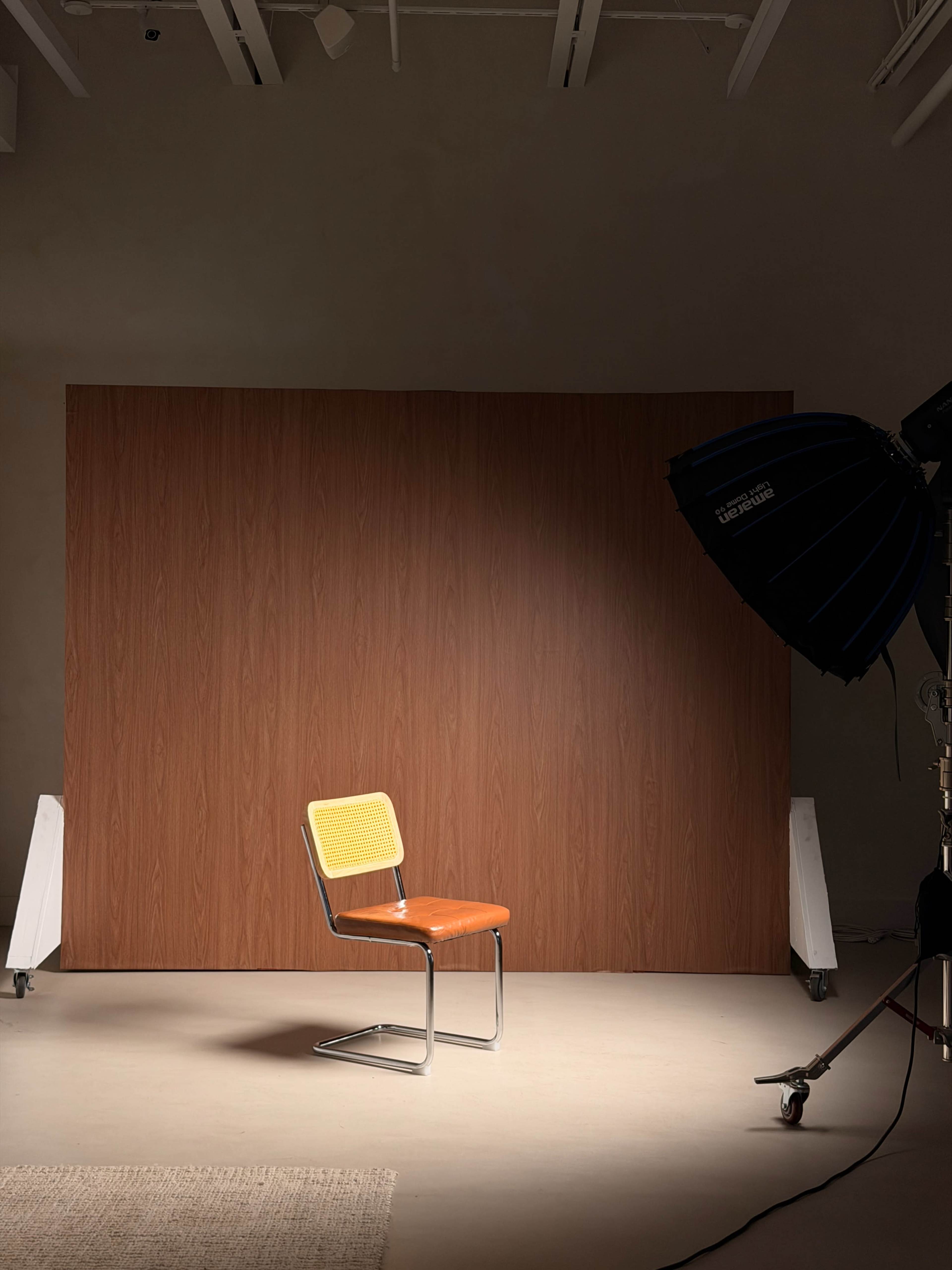 The image shows a single chair with an orange seat set against a wooden panel background, illuminated by a studio lighting setup.