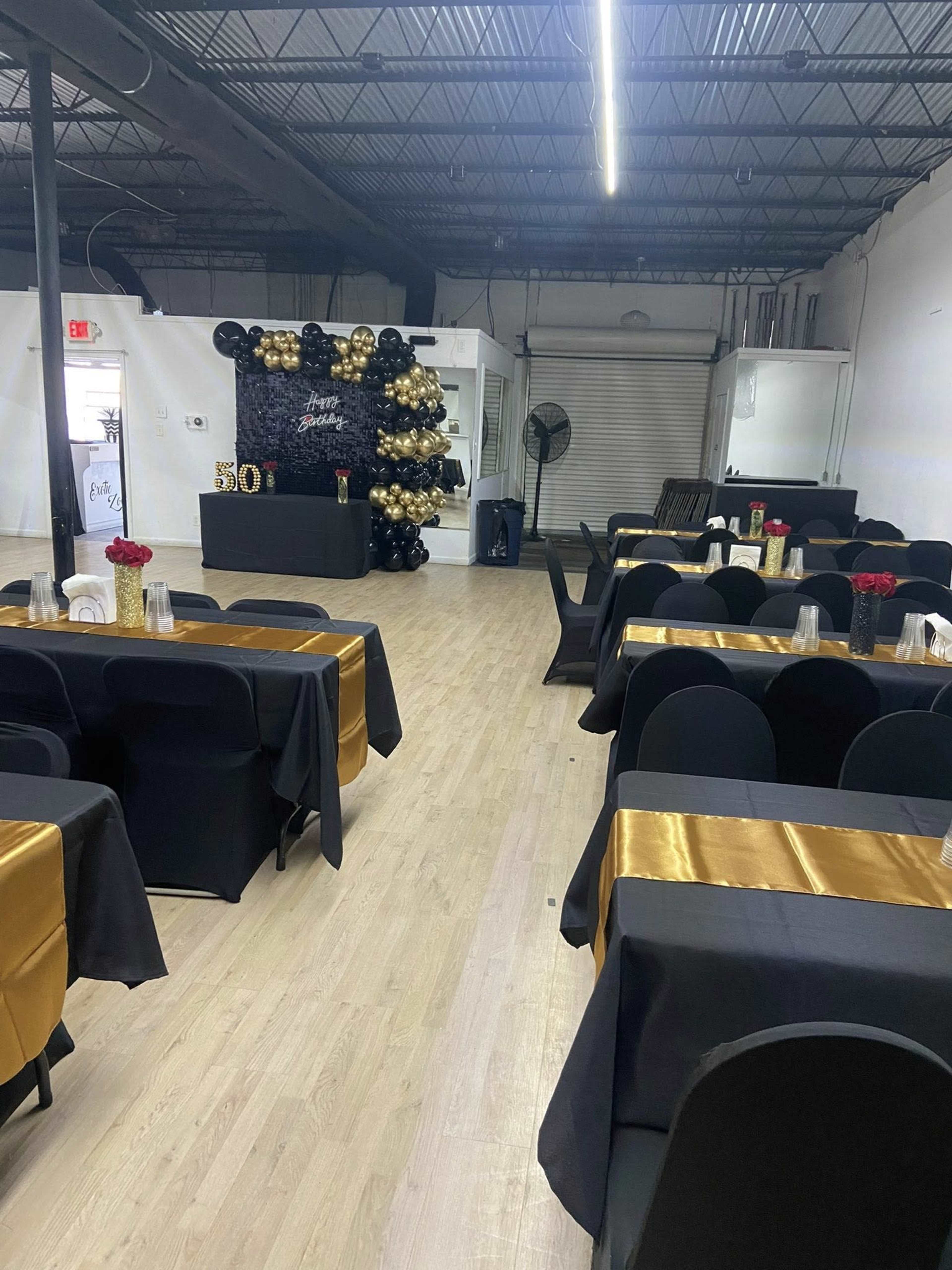 The image shows a spacious event venue set up with tables covered in black and gold tablecloths, arranged in rows, with a decorative wall of balloons and a fan in the background.