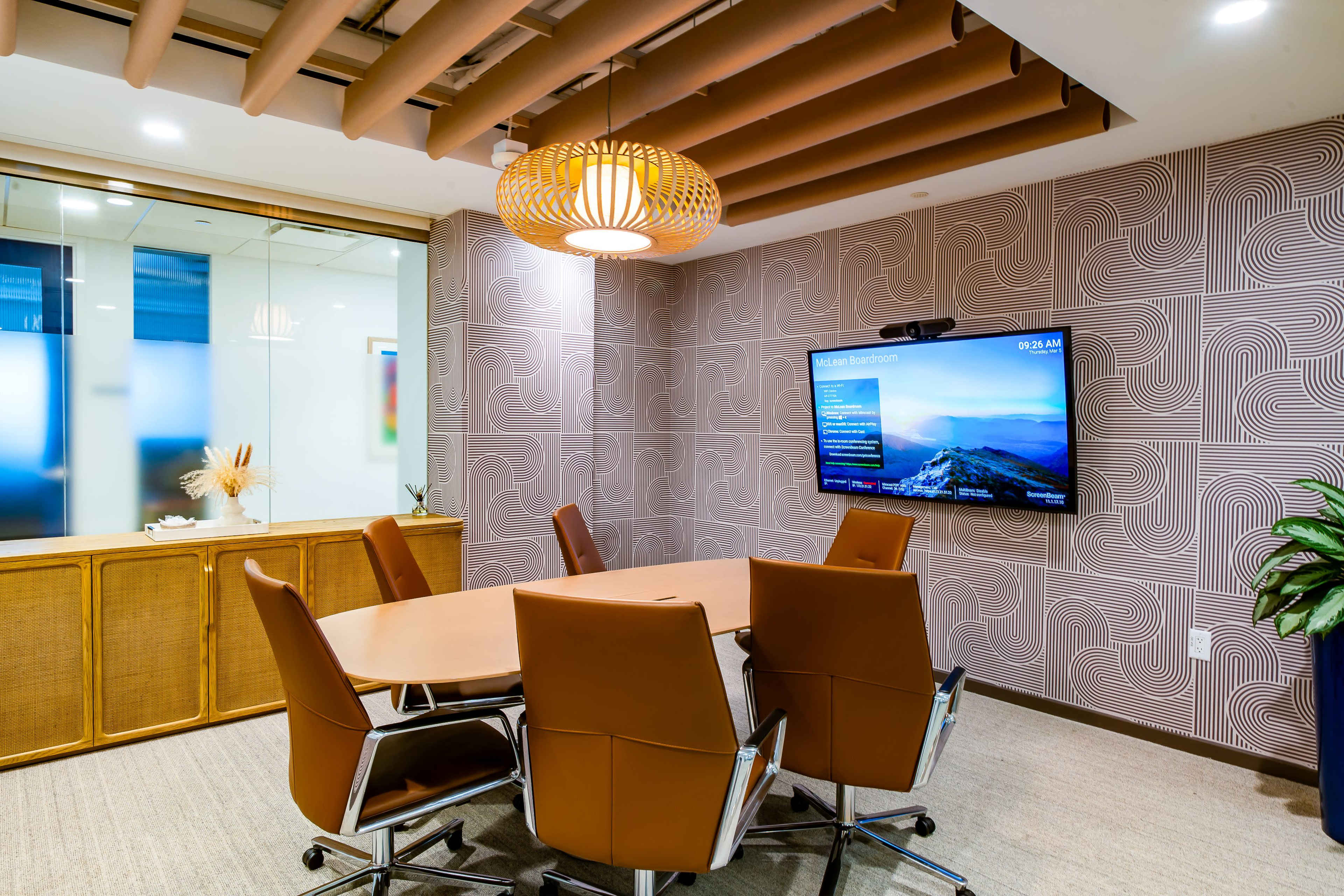 The image shows a modern conference room with a large table, five brown leather chairs, and a wall-mounted screen displaying information.