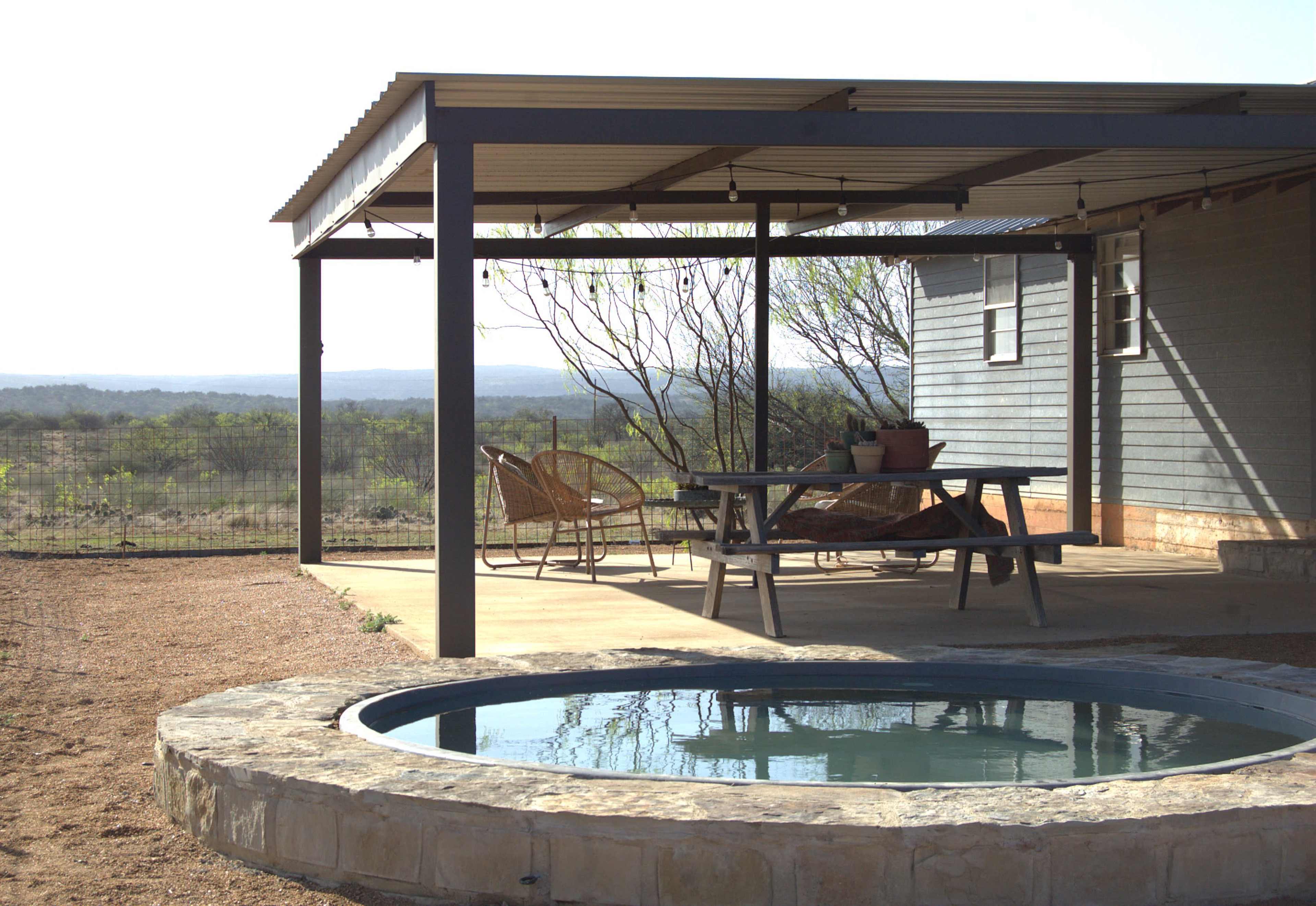 A covered patio with a table and chairs overlooks a small pool and a scenic view of rolling hills.