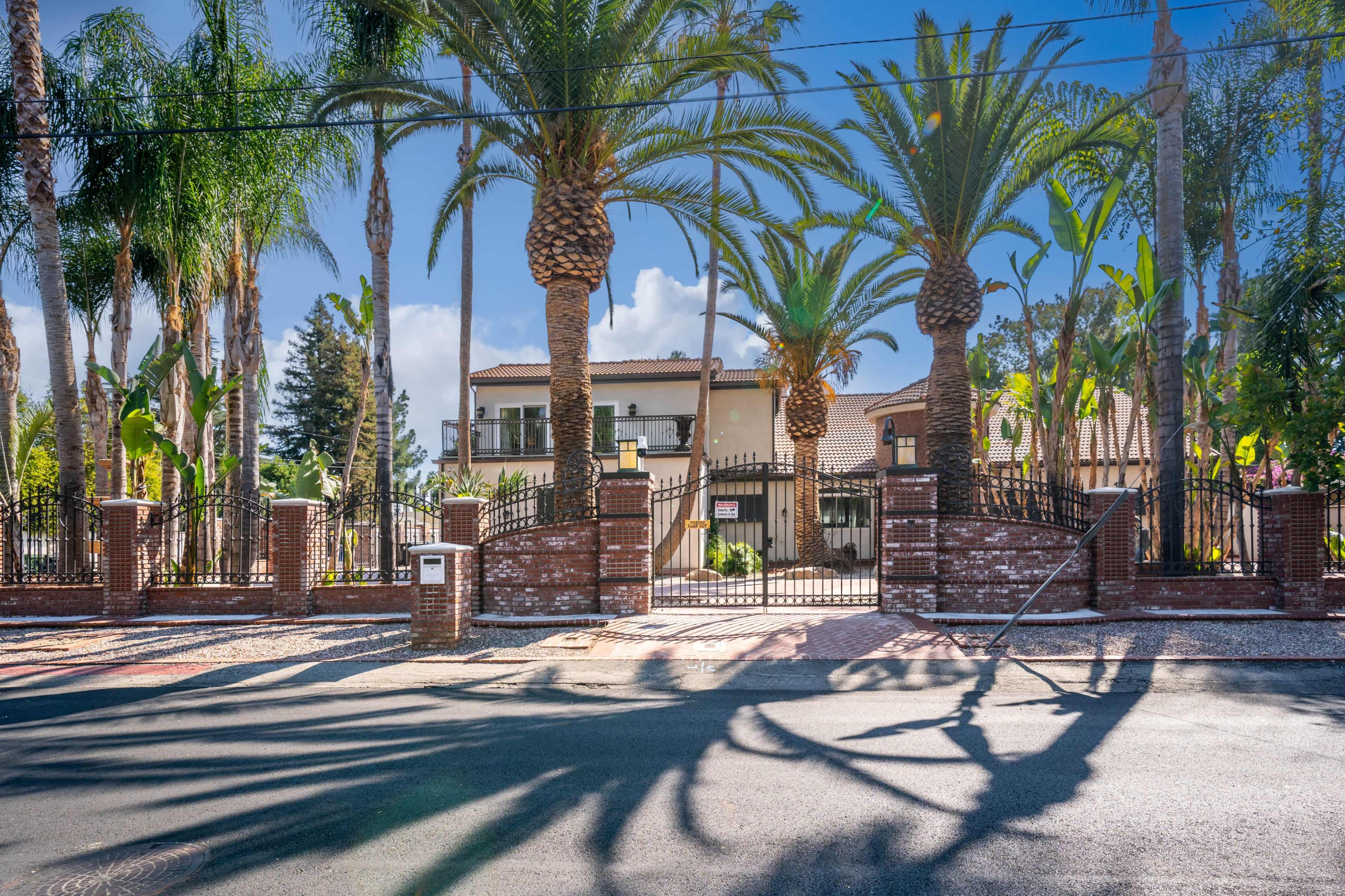 A large, gated entrance with palm trees and a brick wall leads to a two-story house in a residential area.