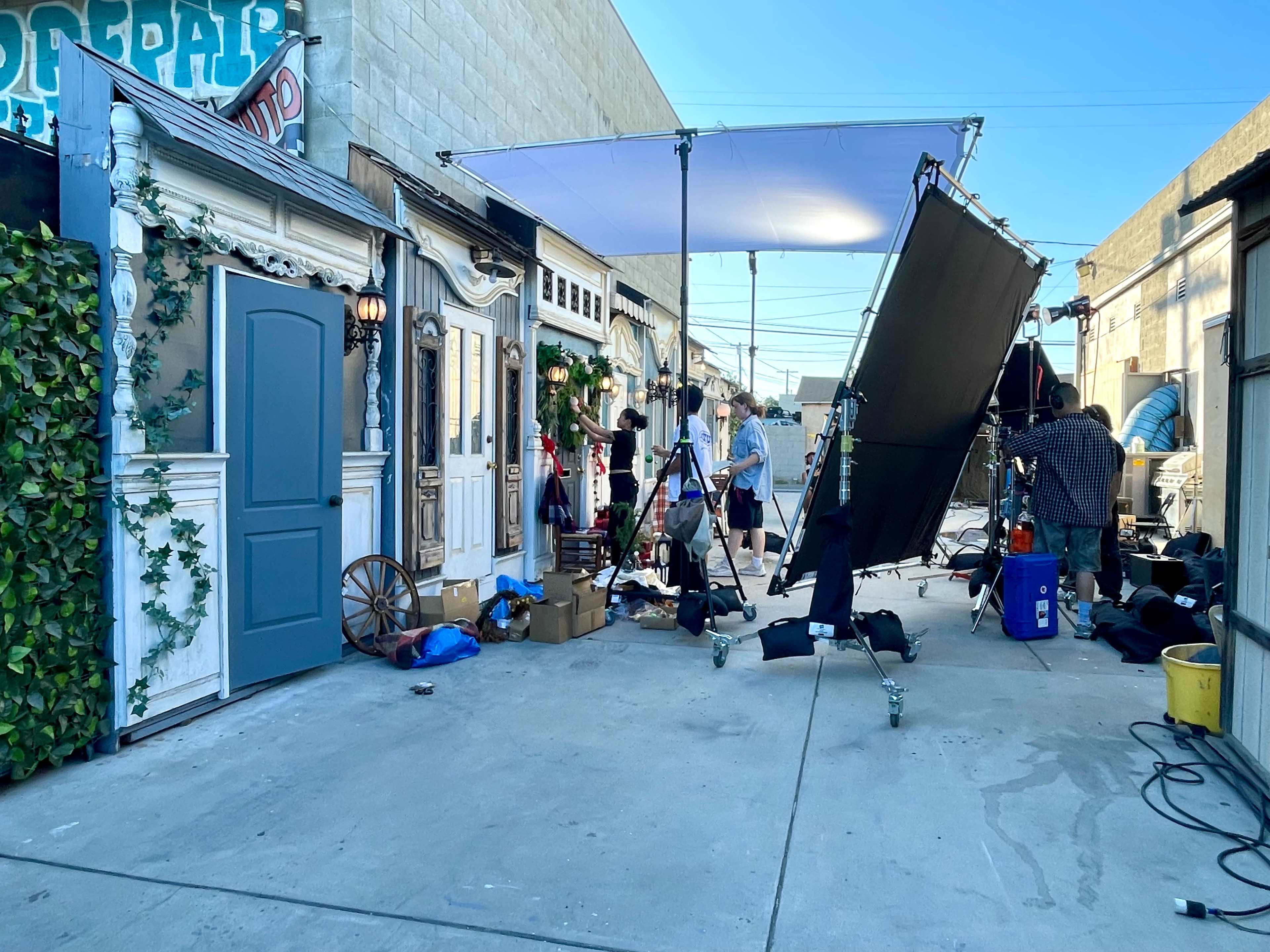 A film set featuring multiple structures, lighting equipment, and crew members preparing for a shoot in an outdoor alleyway.
