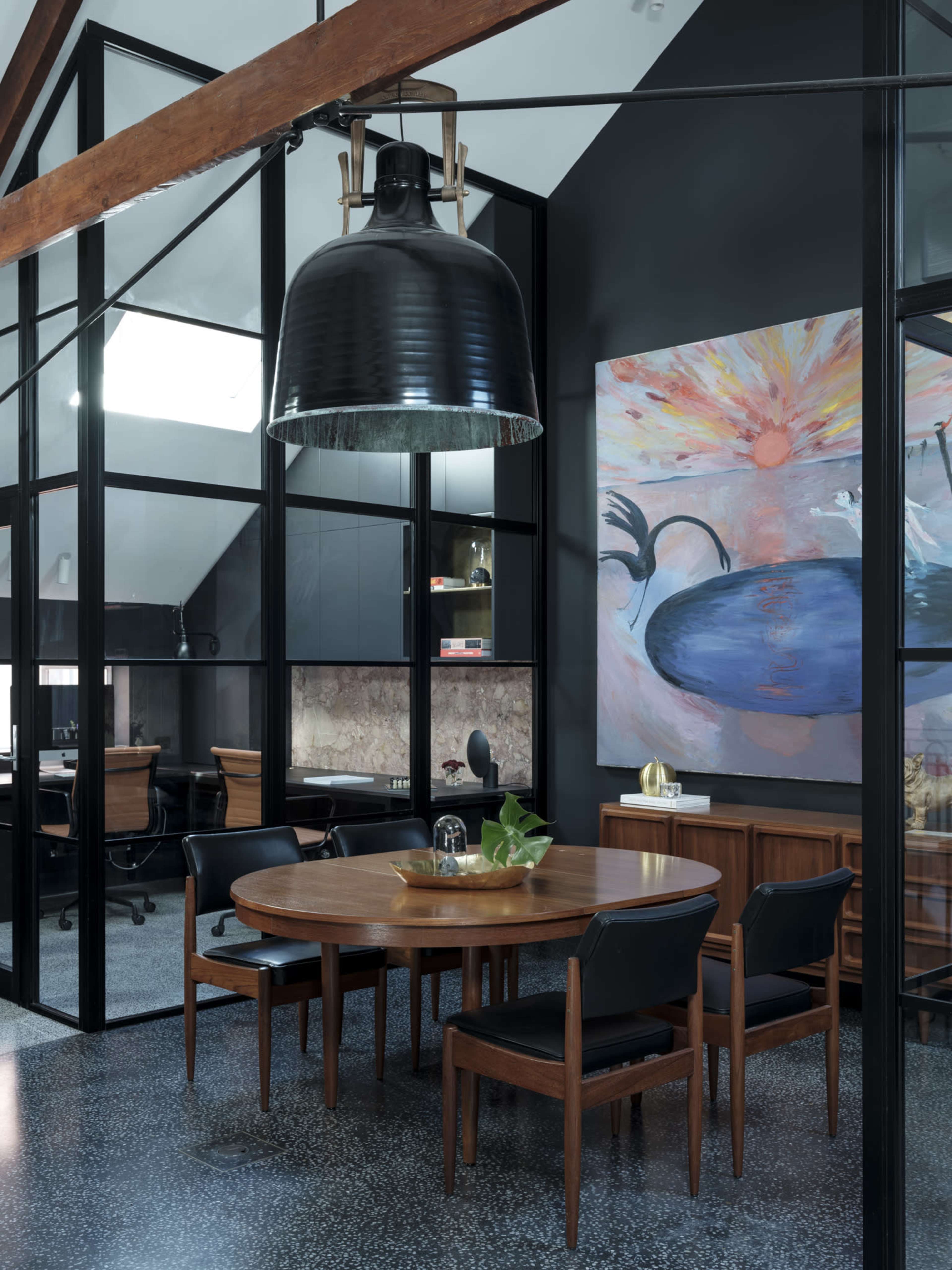 The image shows a modern dining area with a large round wooden table surrounded by black chairs, featuring an industrial-style pendant lamp and a colorful abstract painting on the wall.