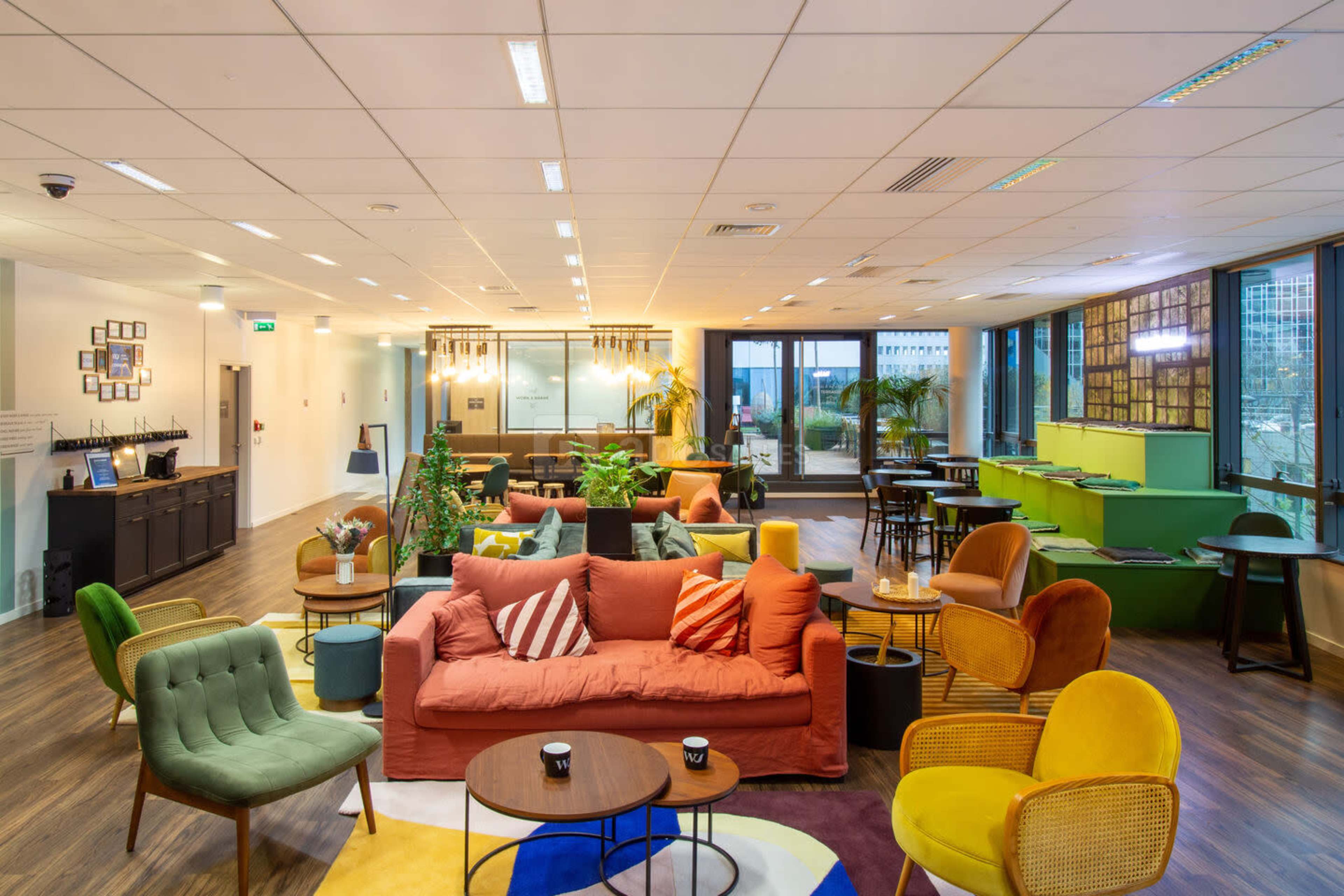 A modern office lounge area featuring a variety of seating arrangements, plants, and large windows that allow natural light to enter.