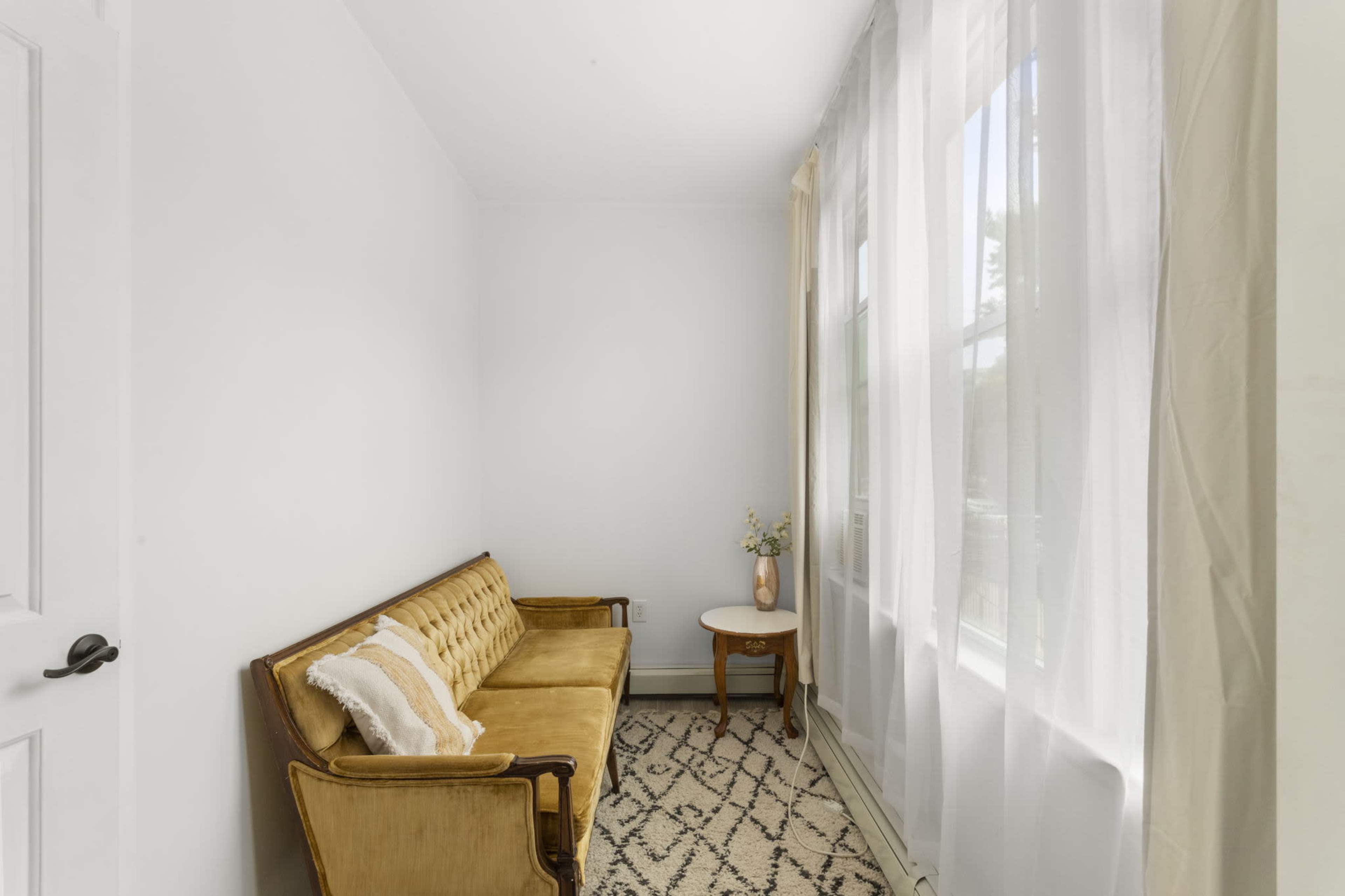 A small room features a vintage yellow couch, a round side table with a vase, and large windows covered with sheer white curtains.