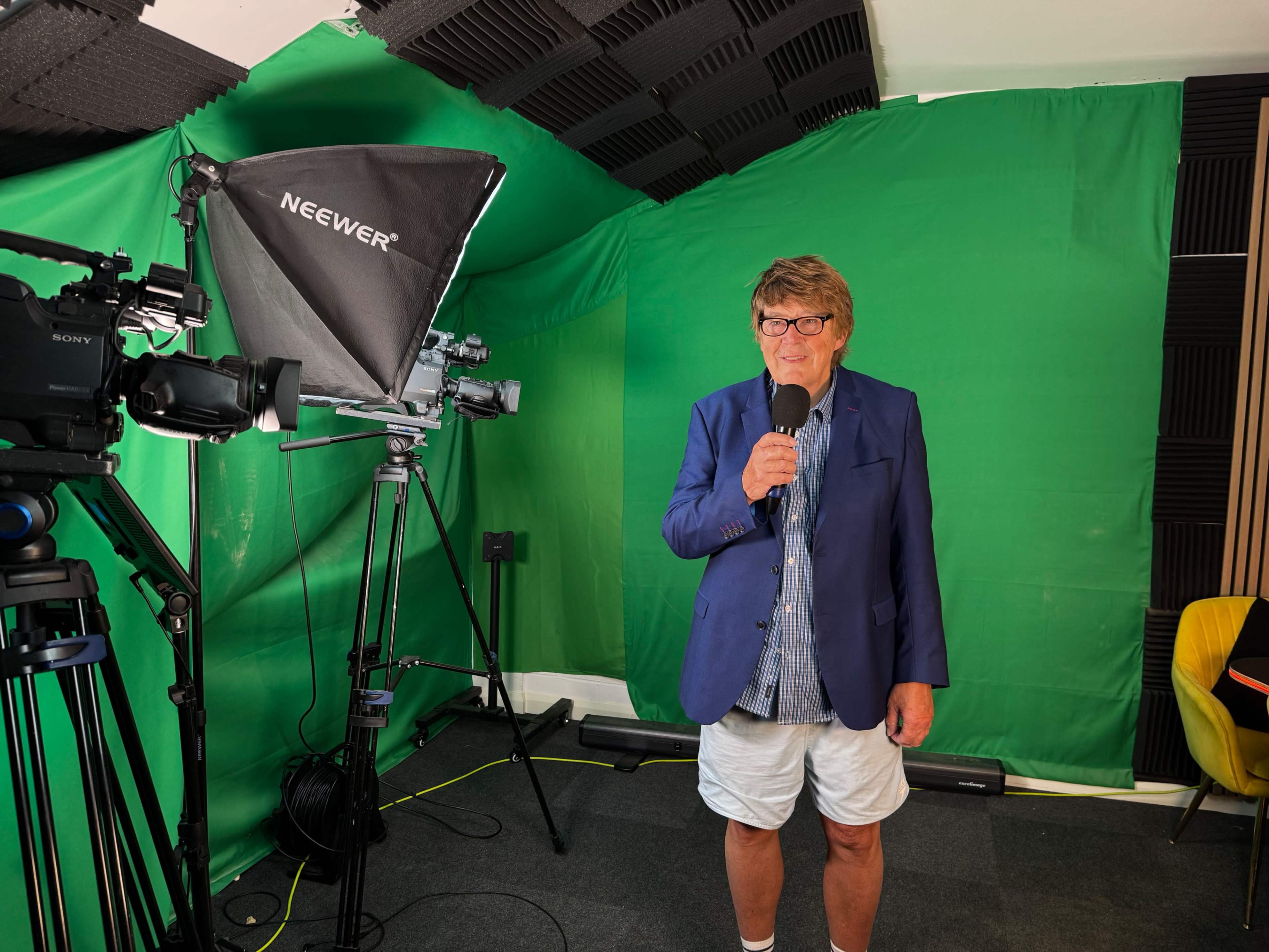 A person stands in front of a green screen, holding a microphone, with cameras and lighting equipment arranged around them.