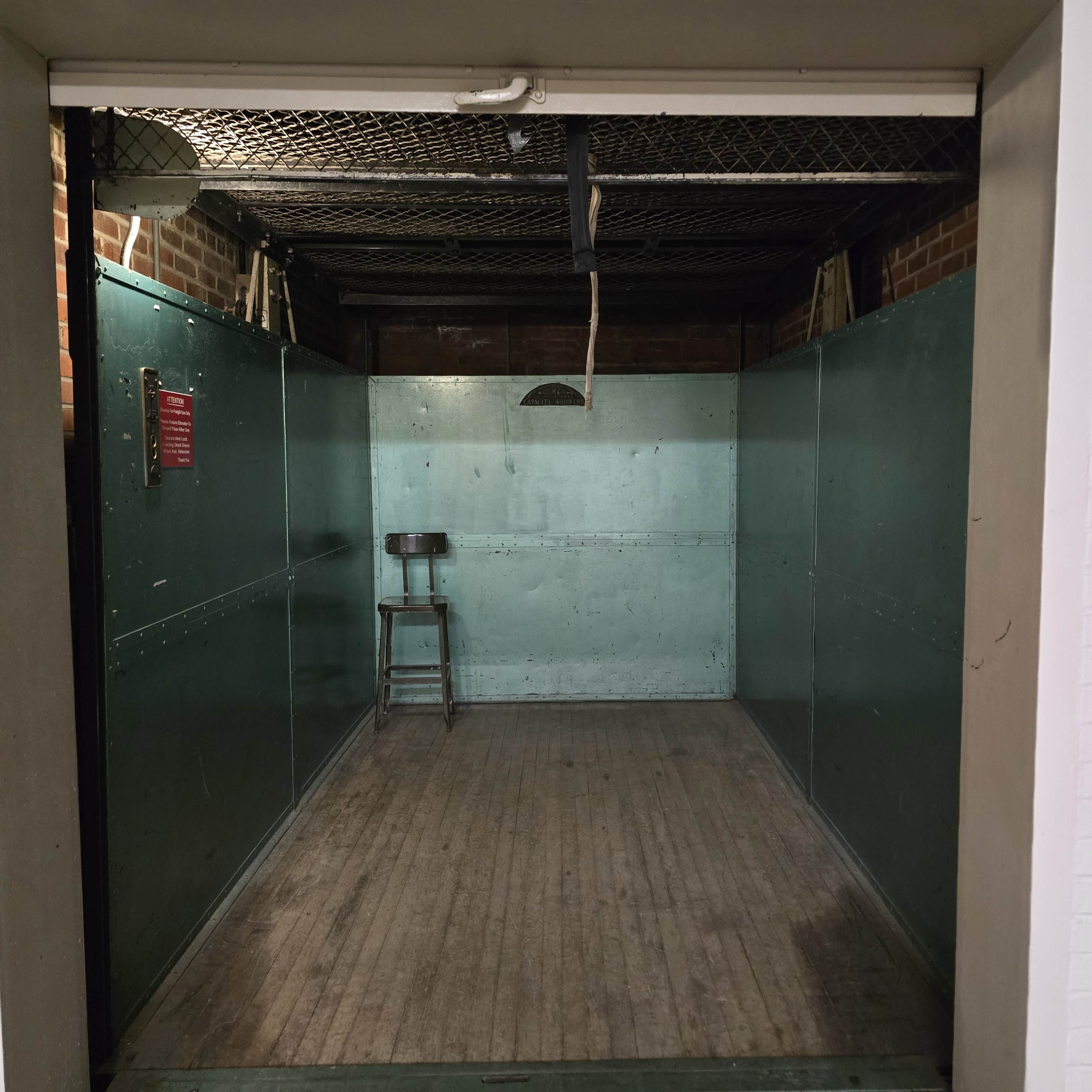 The image shows an empty, industrial-style freight elevator with metal walls and a wooden floor, featuring a small stool in one corner.