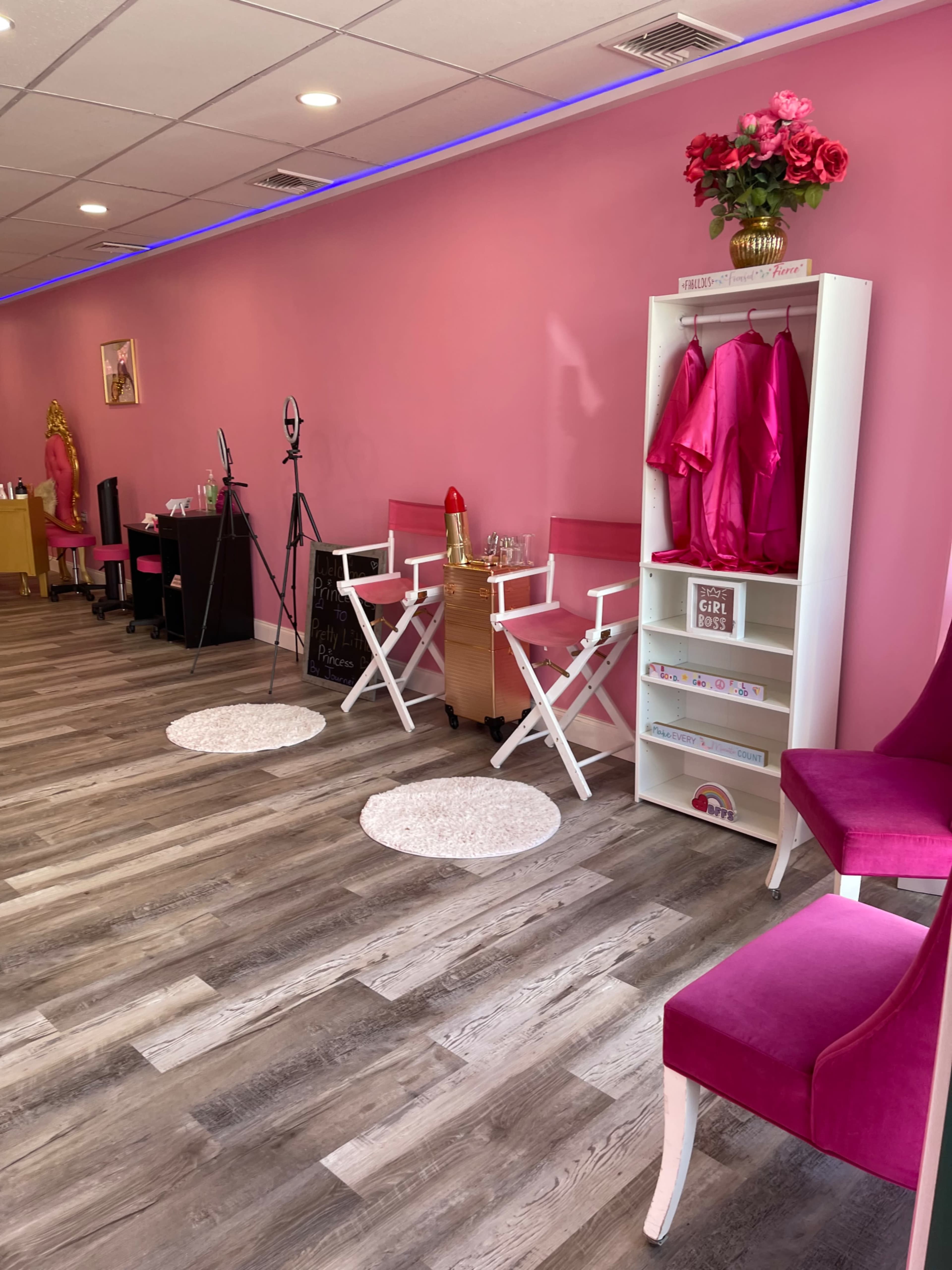 A brightly lit interior of a beauty studio features pink walls, wooden flooring, and styling chairs alongside a rack with pink garments and decor.