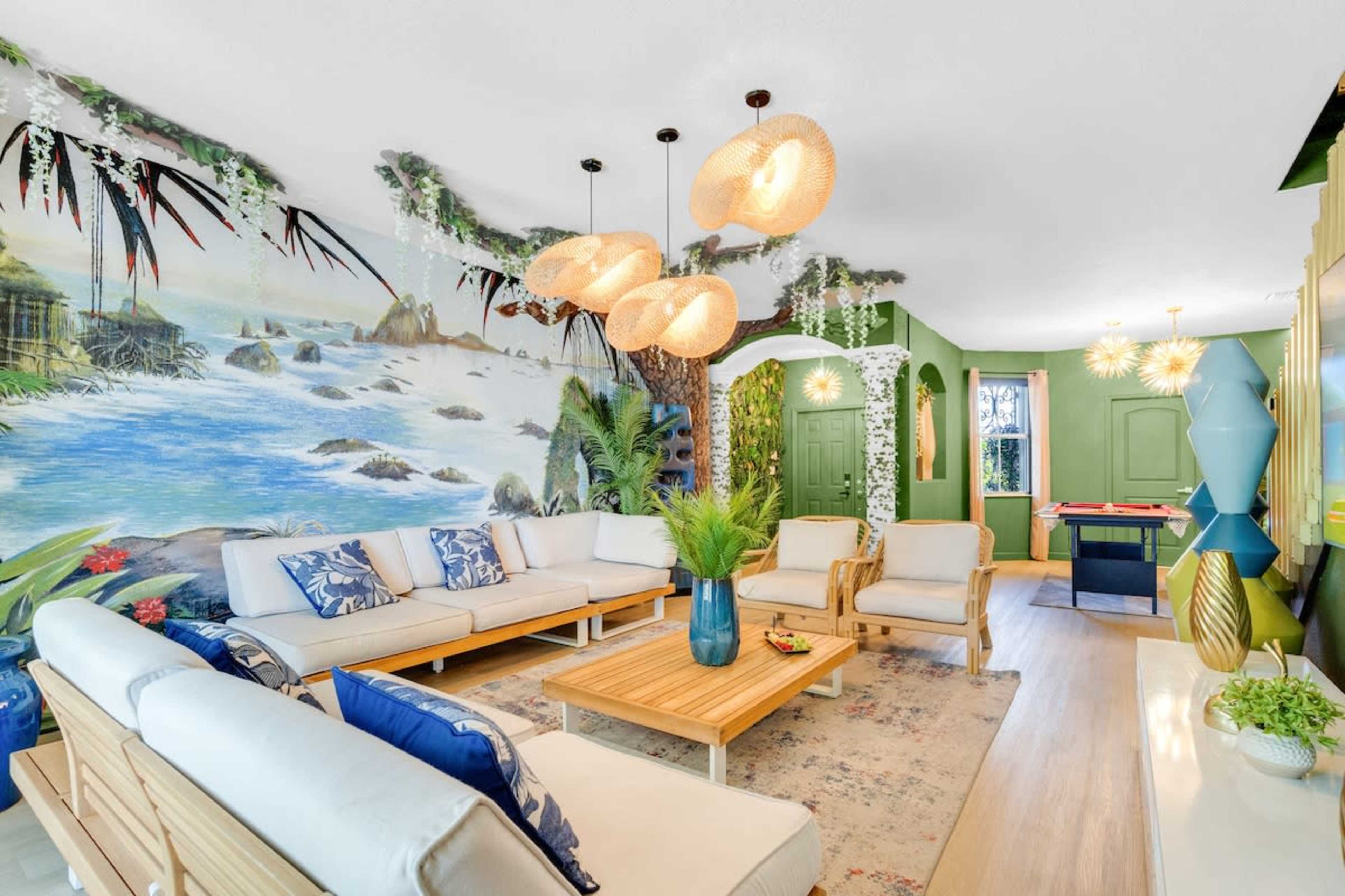 The living room features a mural of a tropical landscape, light-colored furniture, a wooden coffee table, and decorative lighting fixtures overhead.