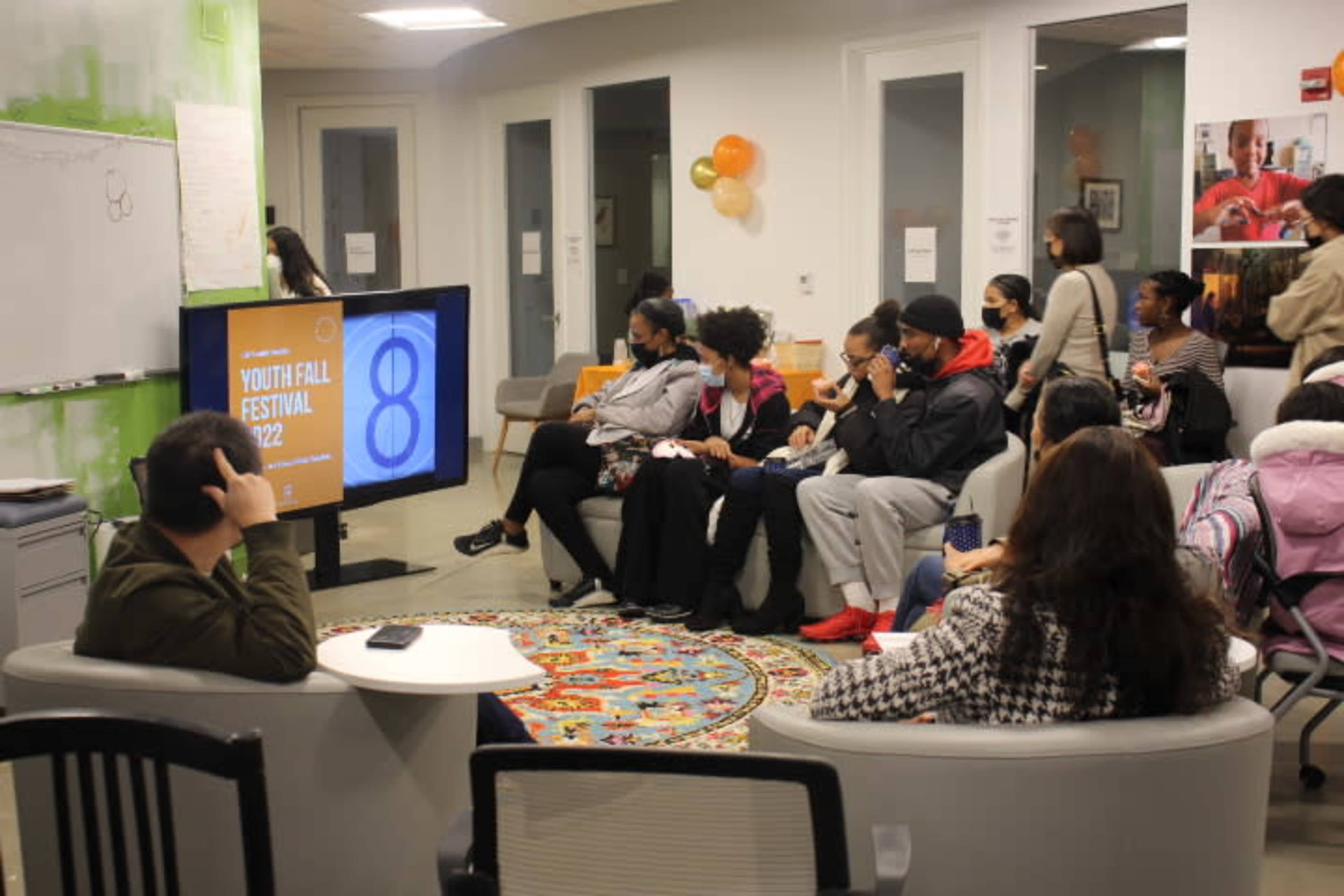 A group of young people sits in a lounge area, watching a presentation titled "Youth Fall Festival 2022" displayed on a television screen.