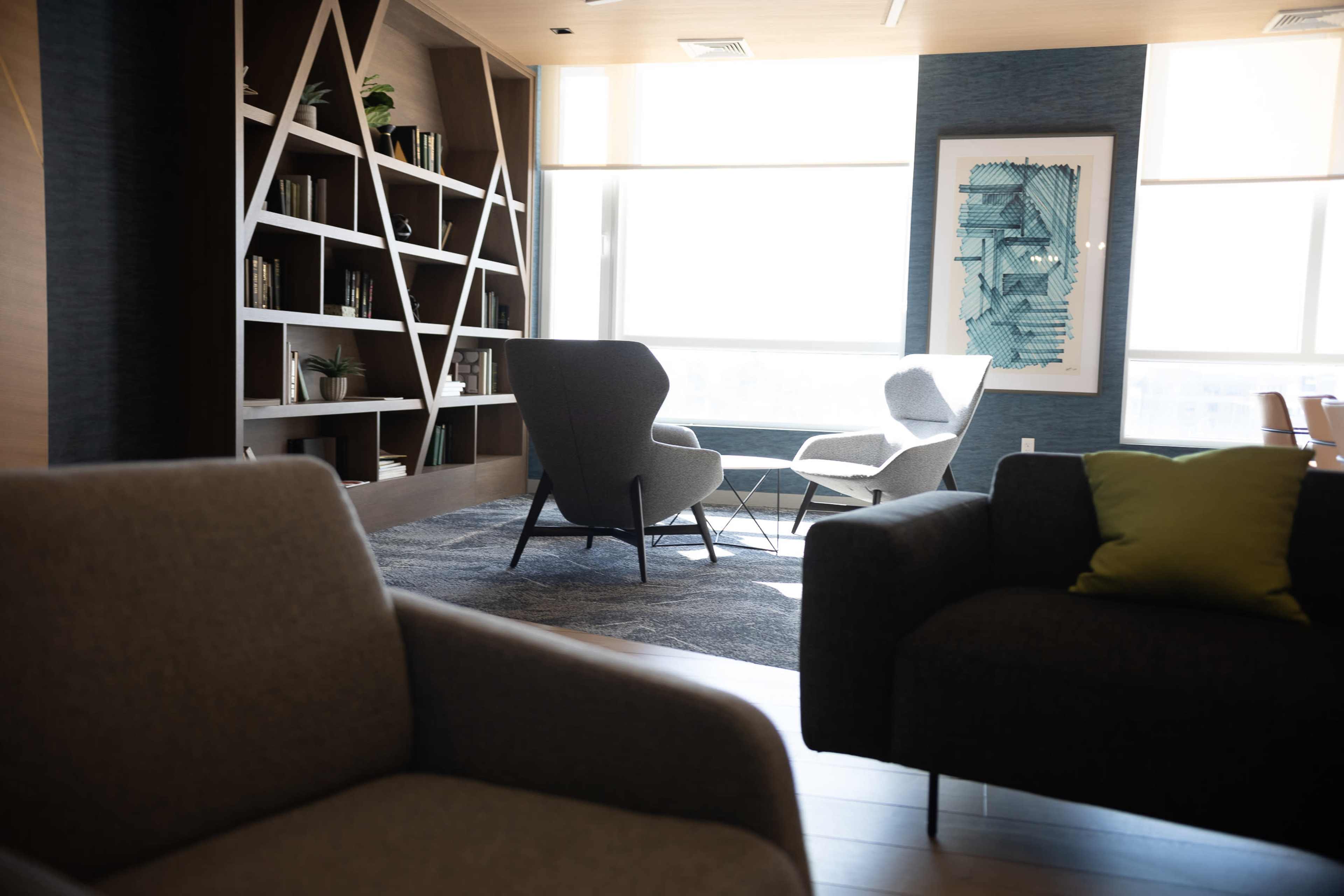 The image shows a modern lounge area with two armchairs and a bookshelf against a wall, illuminated by natural light from large windows.