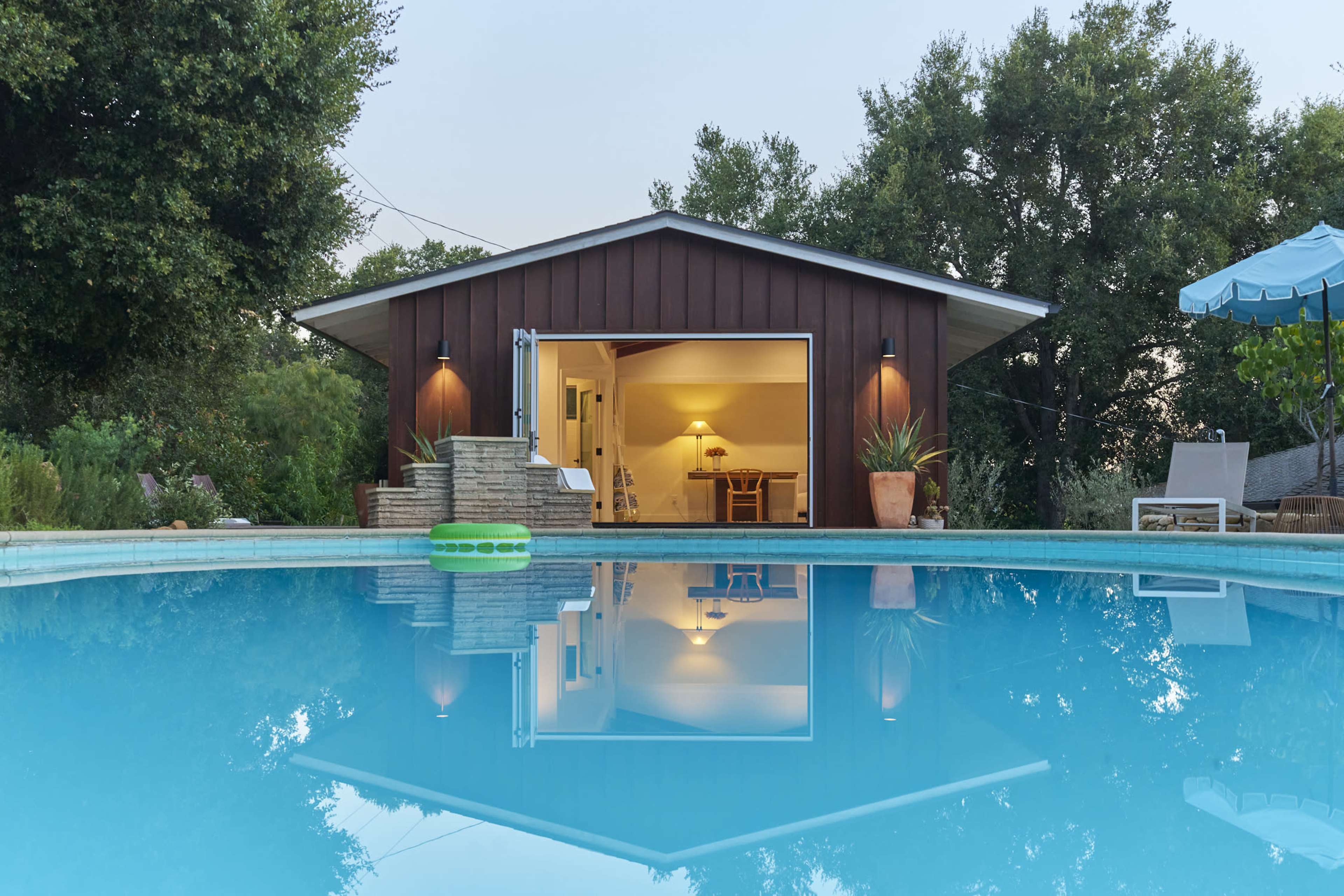 A tranquil backyard scene features a swimming pool reflecting a wooden house with an open door, surrounded by lush greenery.