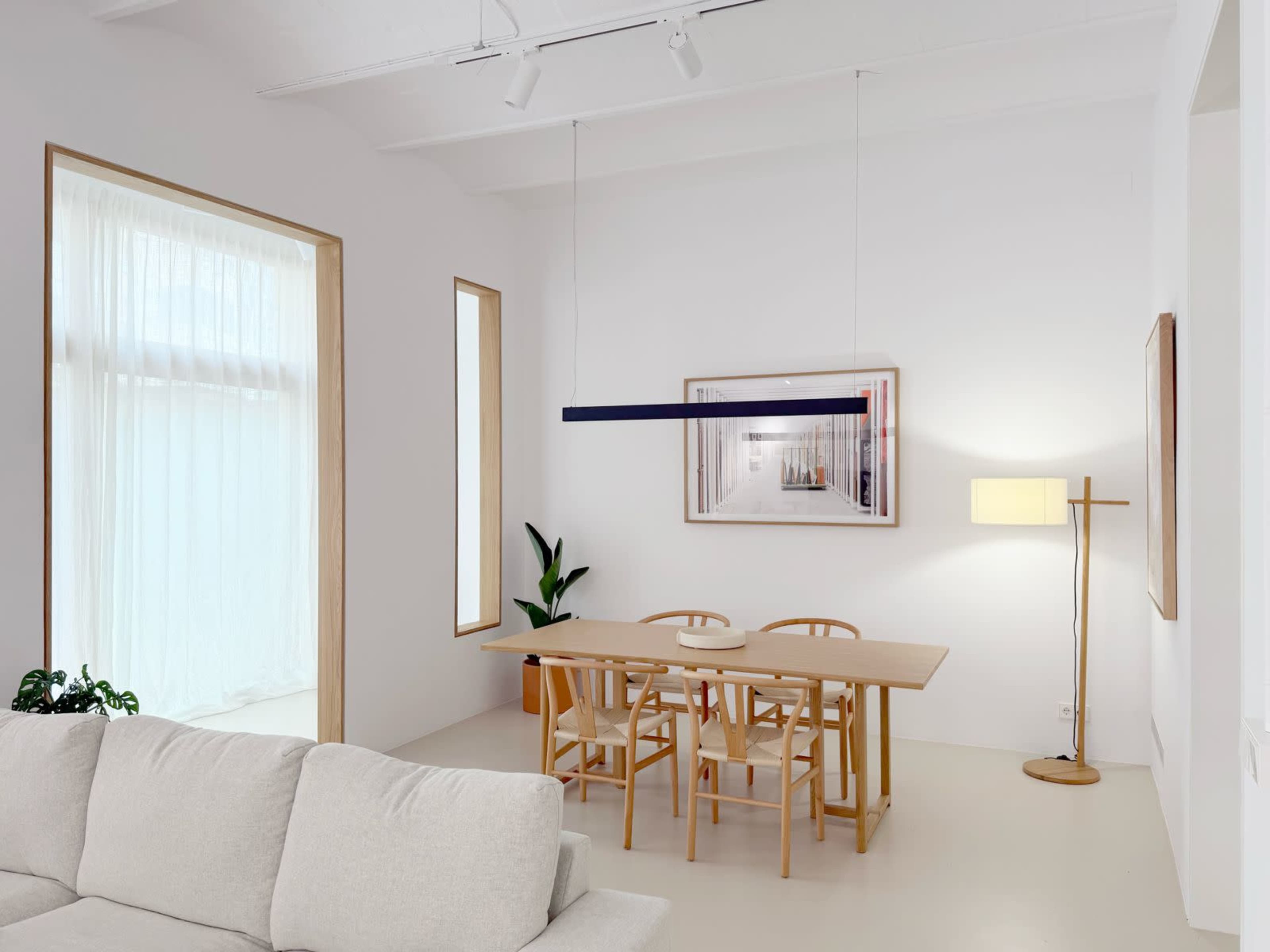 The image shows a minimalist dining area with a wooden table surrounded by chairs, illuminated by a modern light fixture, and featuring large windows with sheer curtains.