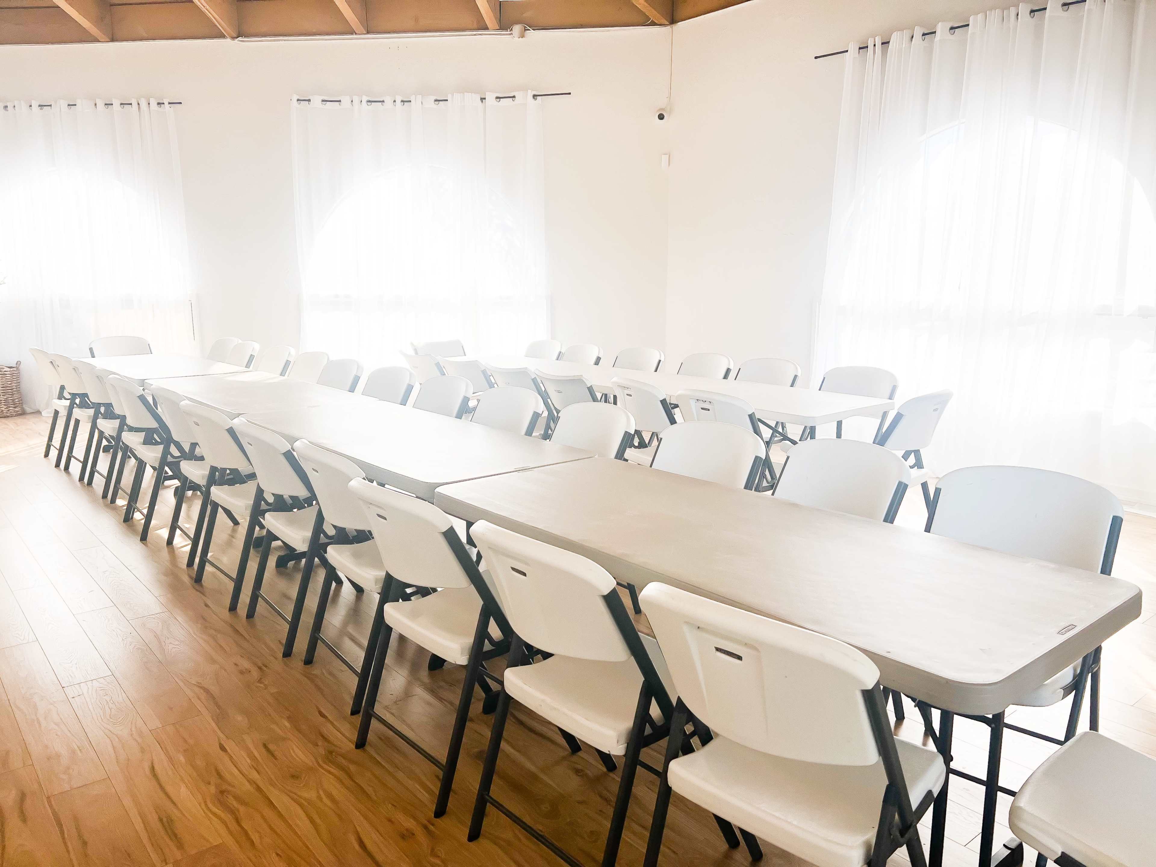 A spacious room arranged with several folding tables and chairs set up for a meeting or event, with large windows covered by white curtains allowing natural light to enter.
