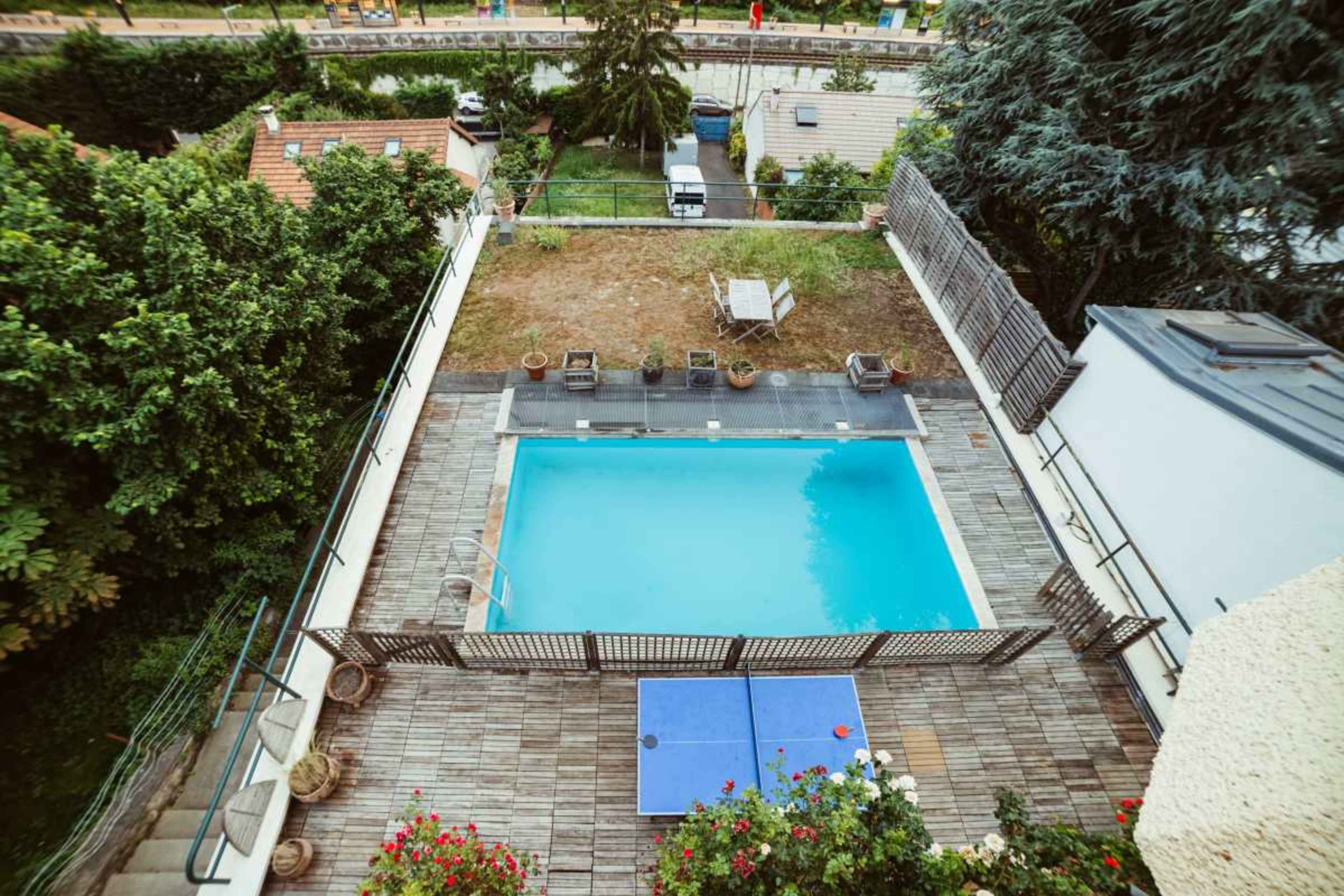 The image shows a rectangular swimming pool surrounded by a wooden deck and greenery, viewed from an elevated perspective.