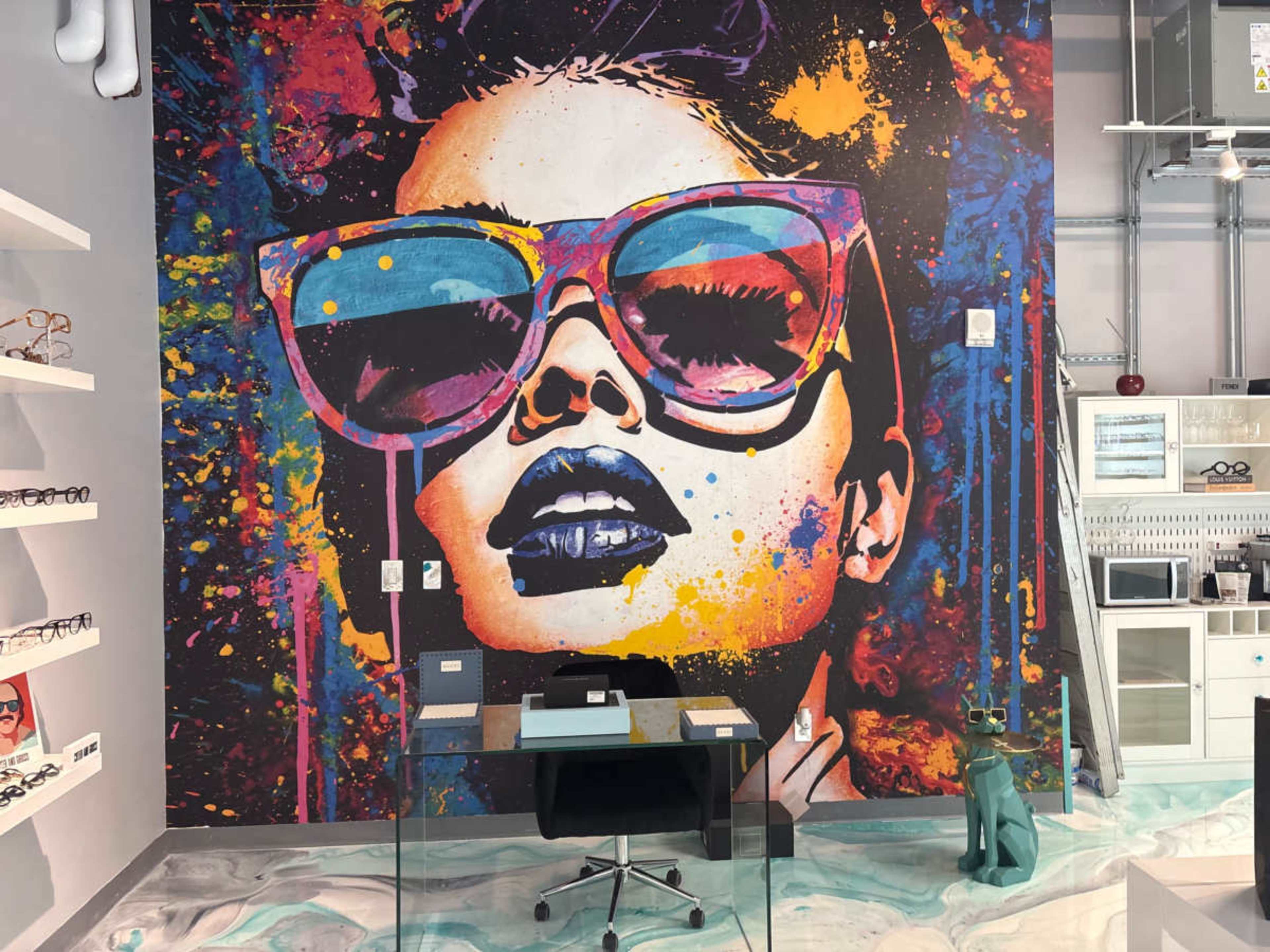 The image shows a vibrant mural of a woman wearing oversized sunglasses, displayed on a wall in a store, with a glass display table in front.