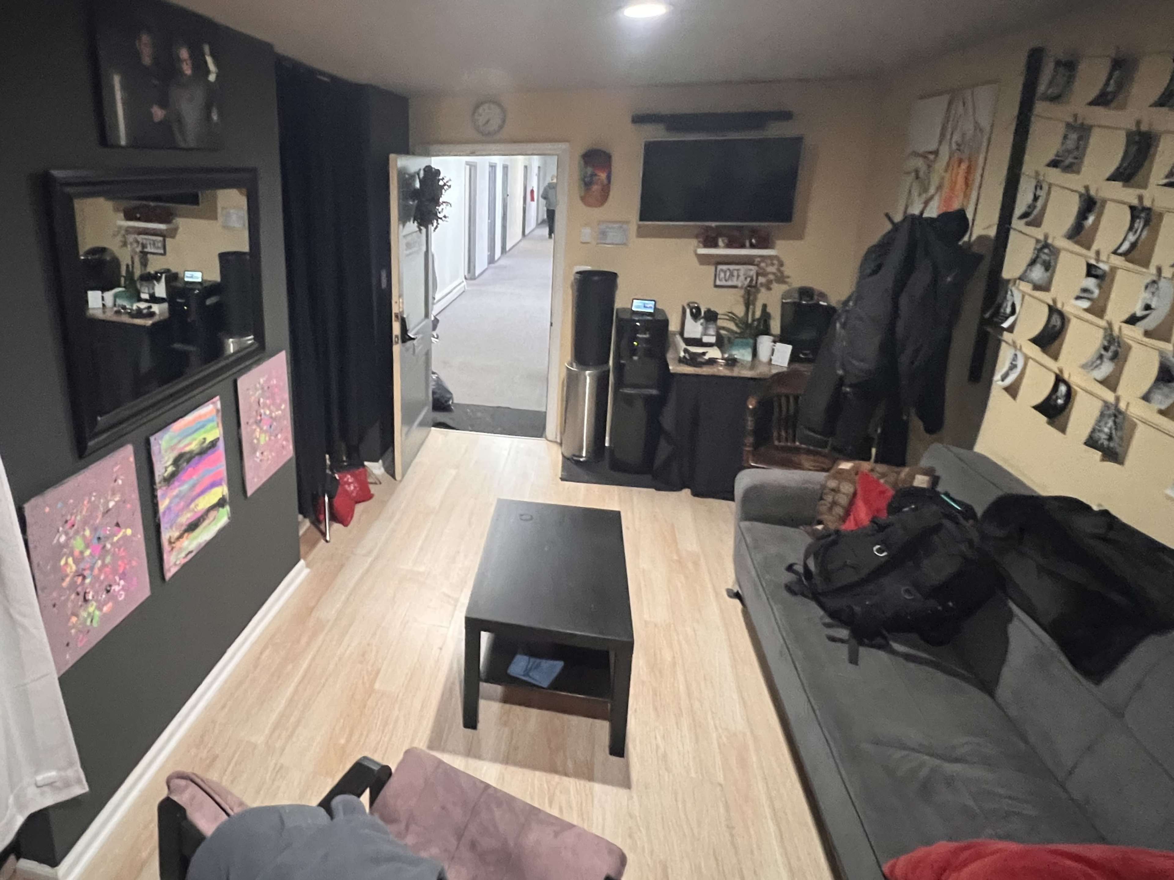 A casual living space with a couch, a coffee table, and various items like bags and a coffee machine, along with wall decorations.