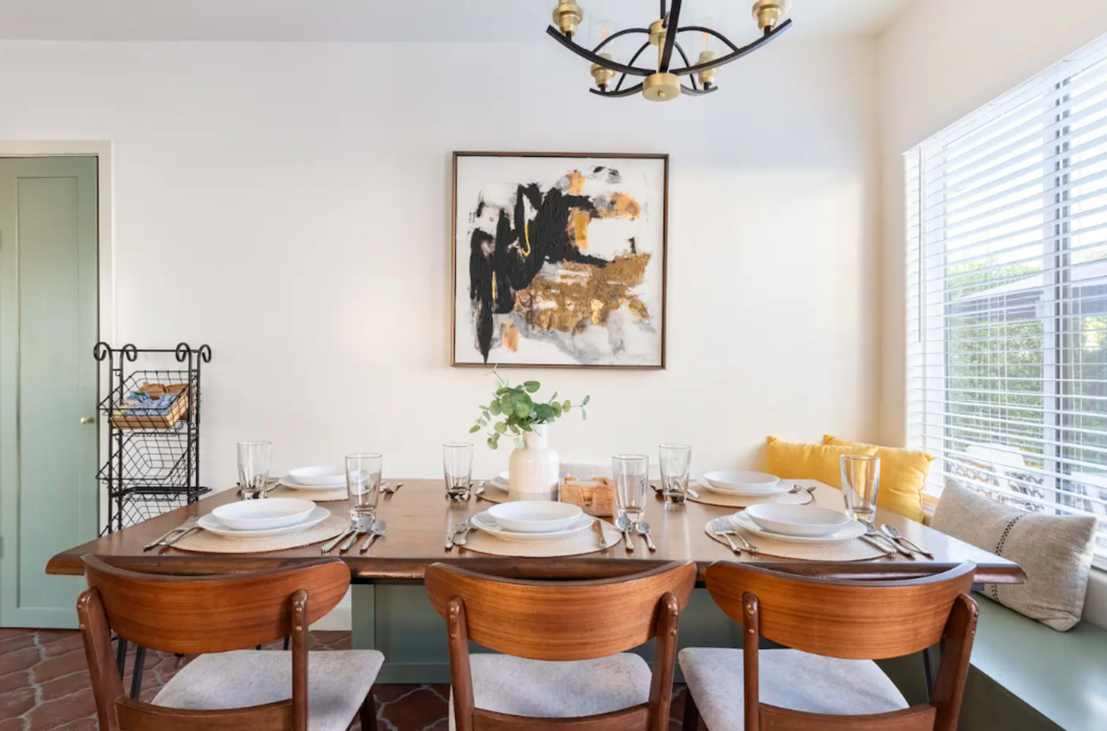 A wooden dining table set for a meal, surrounded by four chairs, with a piece of abstract wall art and a window letting in natural light.