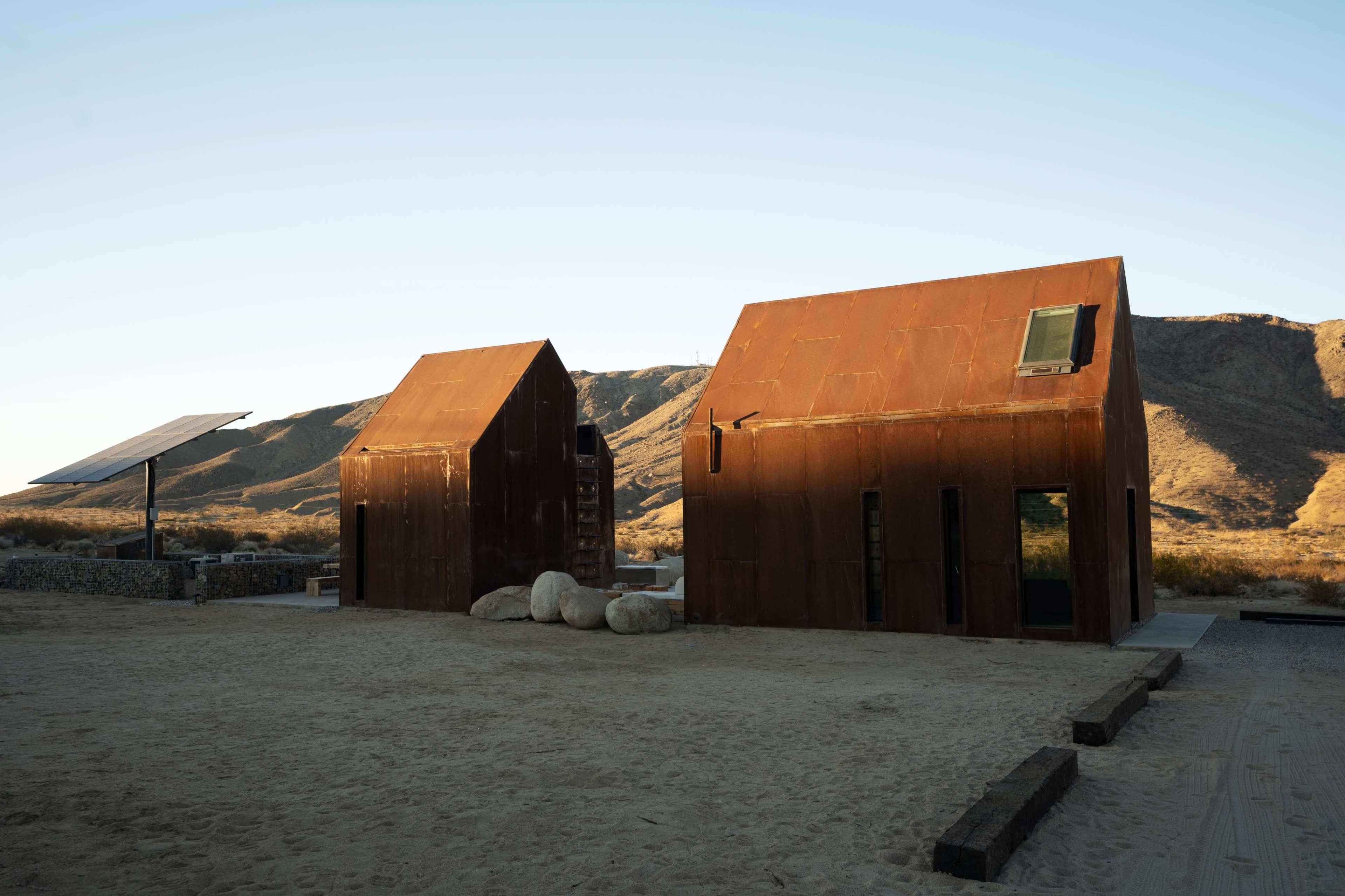 Two rust-colored metal structures with sloped roofs sit in a sandy area near mountains, alongside a solar panel installation.