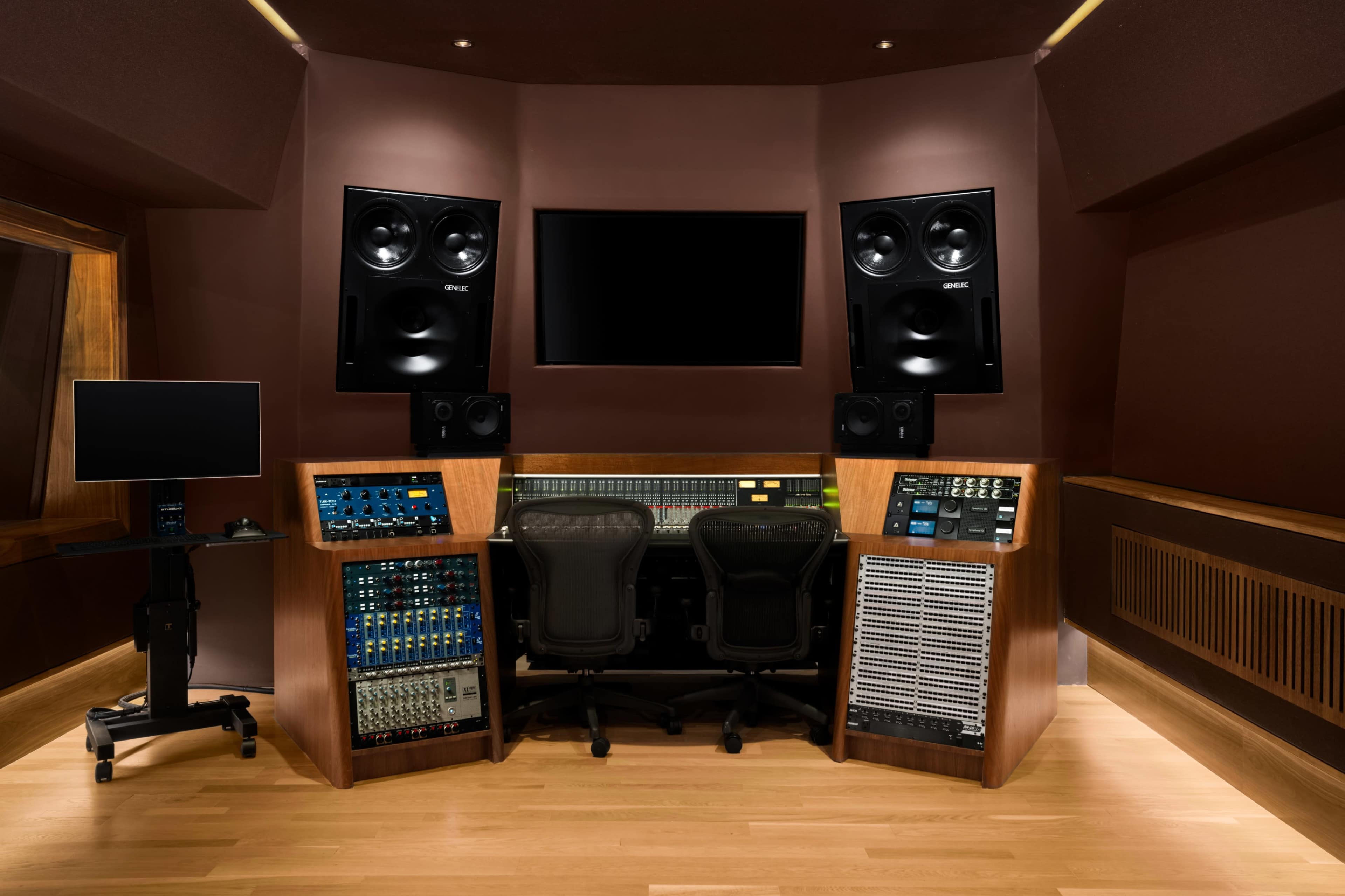 The image shows a recording studio with a console at the center, flanked by large speakers and a monitor, all set in a modern, wood-paneled environment.