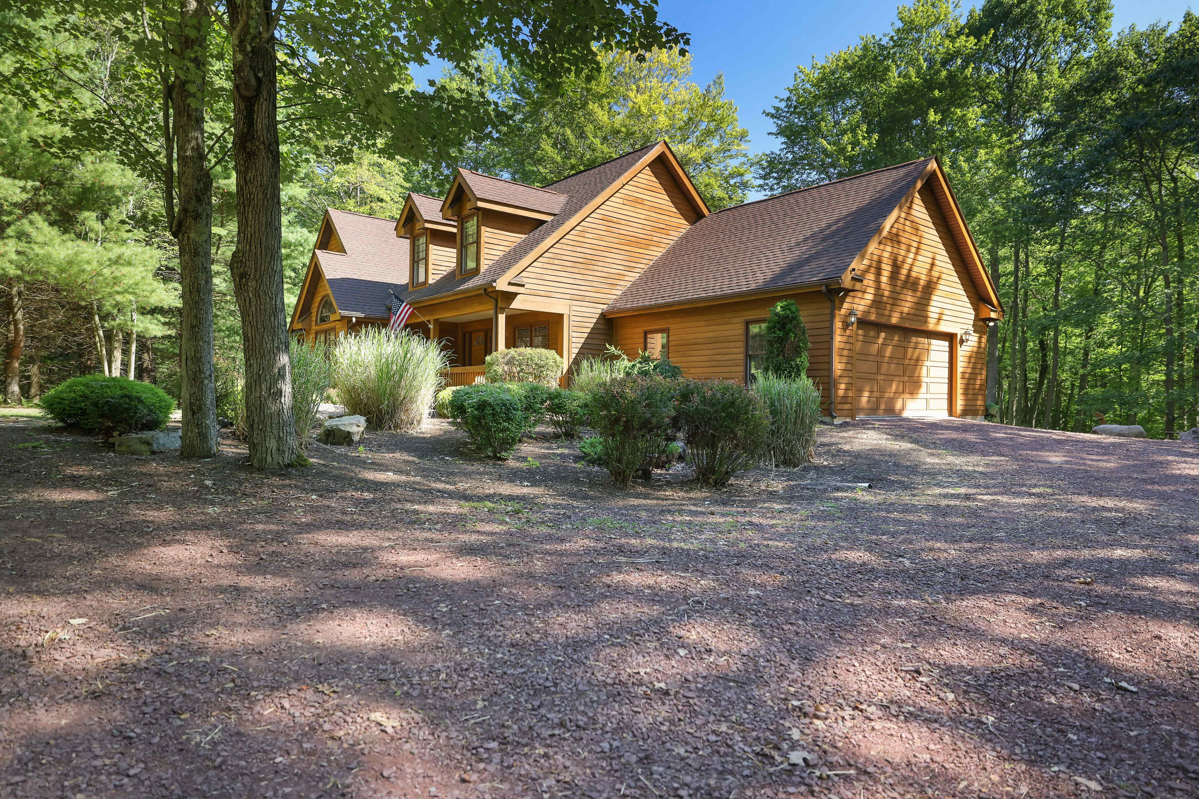 A two-story wooden house with a sloped roof sits on a gravel drive surrounded by trees and shrubs.